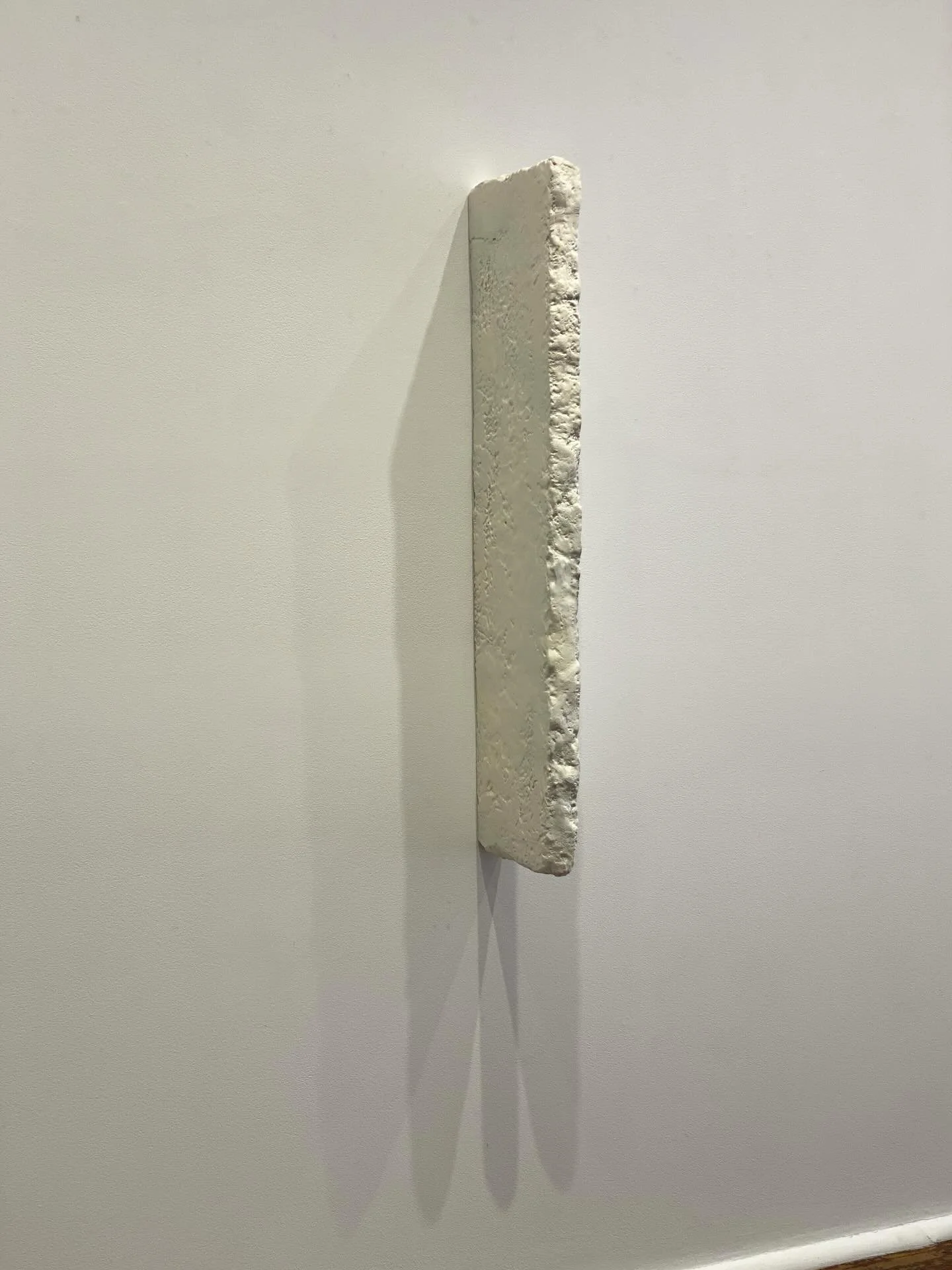 A white rectangular sculpture with rough edges, mounted on a white wall, casting a shadow.