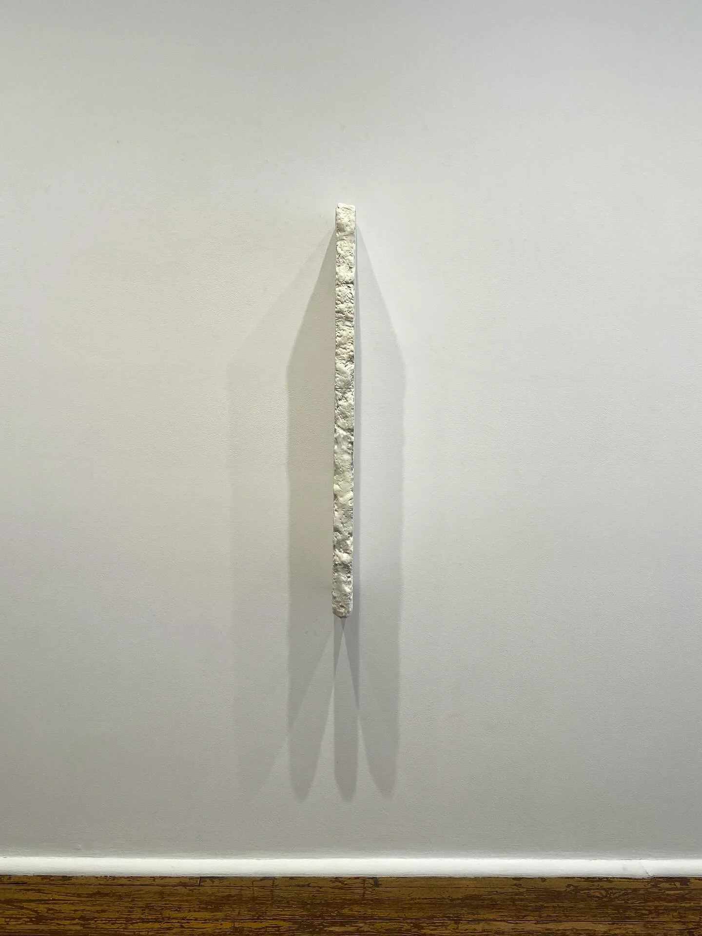 A vertical, textured, white sculpture or object mounted on a plain white wall, casting a shadow to the left.