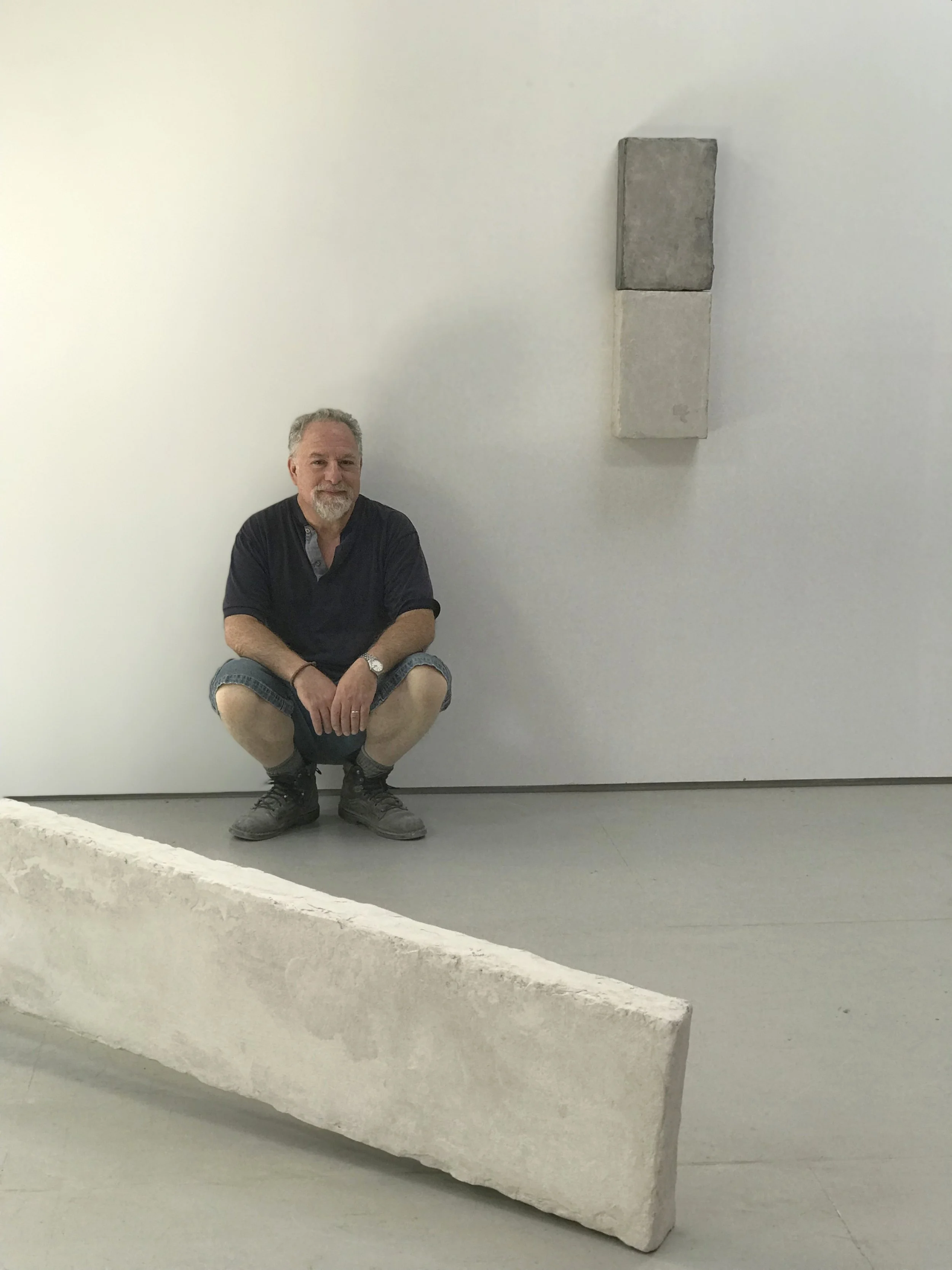 A man crouching in front of a large, rectangular stone sculpture in a gallery or museum setting.