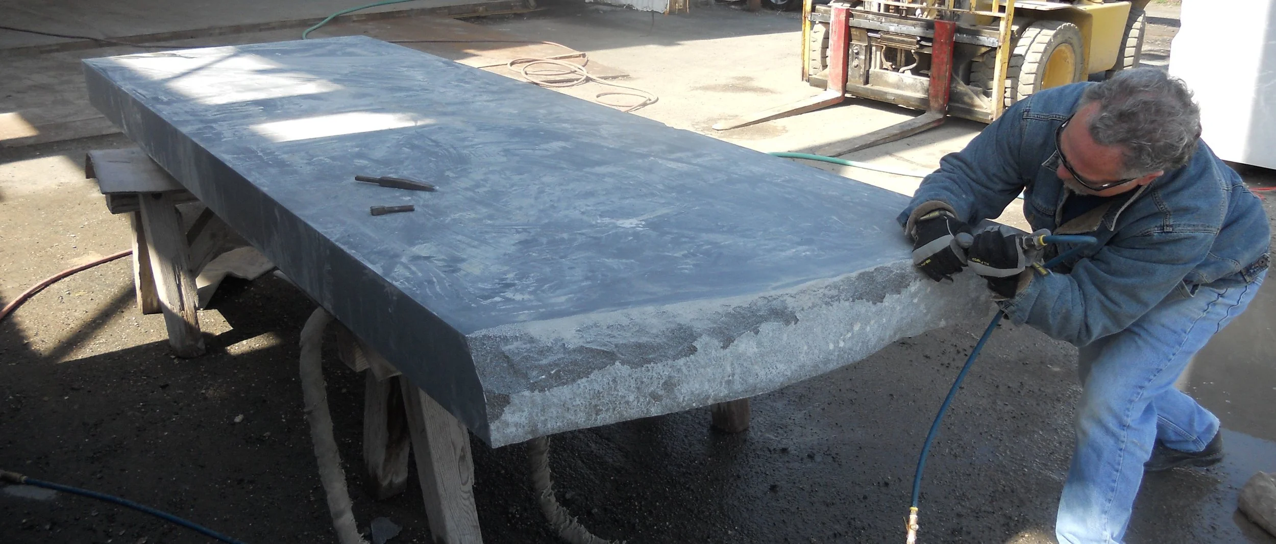 A man wearing glasses, gloves, and a denim jacket is working with a power tool on a large, flat piece of dark stone or granite supported by wooden sawhorses in a workshop or outdoor setting. A forklift and construction equipment are visible in the background.