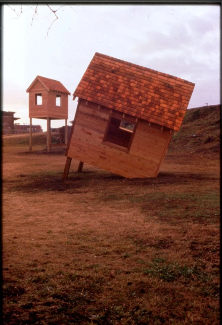 Two small wooden house-shaped structures on stilts, with the larger house tilted at an angle. The scene appears to be an outdoor playground or art installation with a slightly overcast sky.