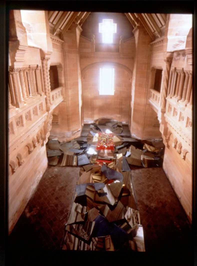 An art installation inside a church-like structure with wooden walls, arched windows, and a high ceiling. The installation features shards of broken mirrors and reflective surfaces on the floor with a colorful pile of objects at the center, illuminat