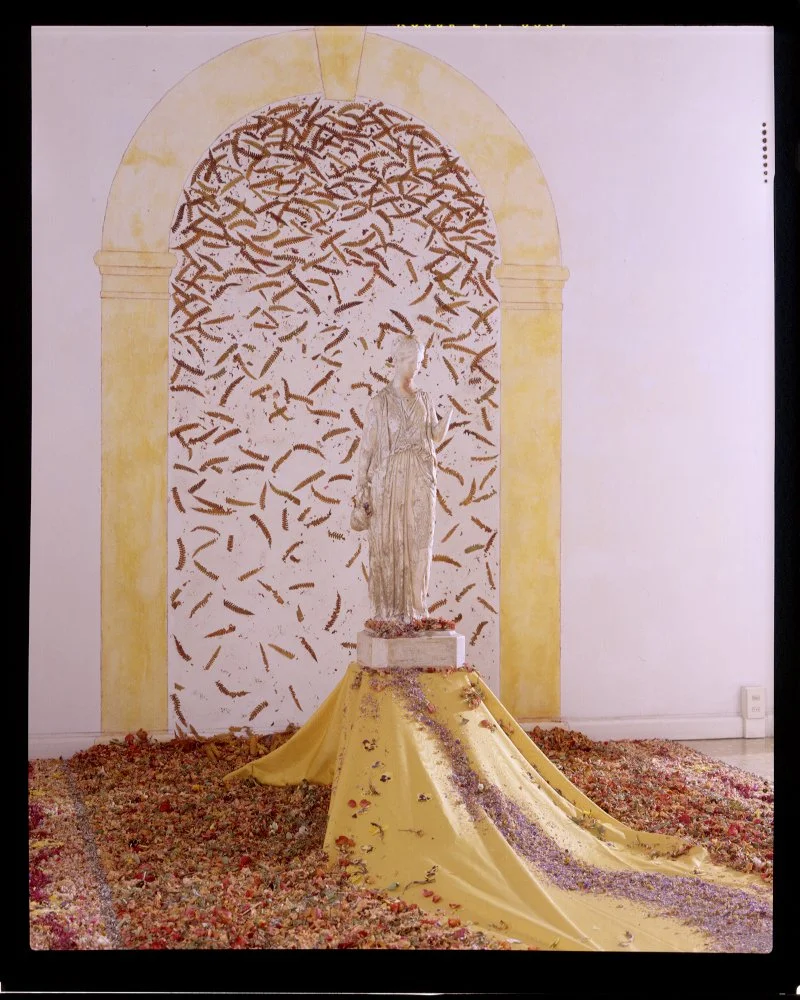 A classical statue of a woman on a pedestal draped with yellow fabric, surrounded by flower petals, with a painted wall background of an arch and abstract brown shapes.