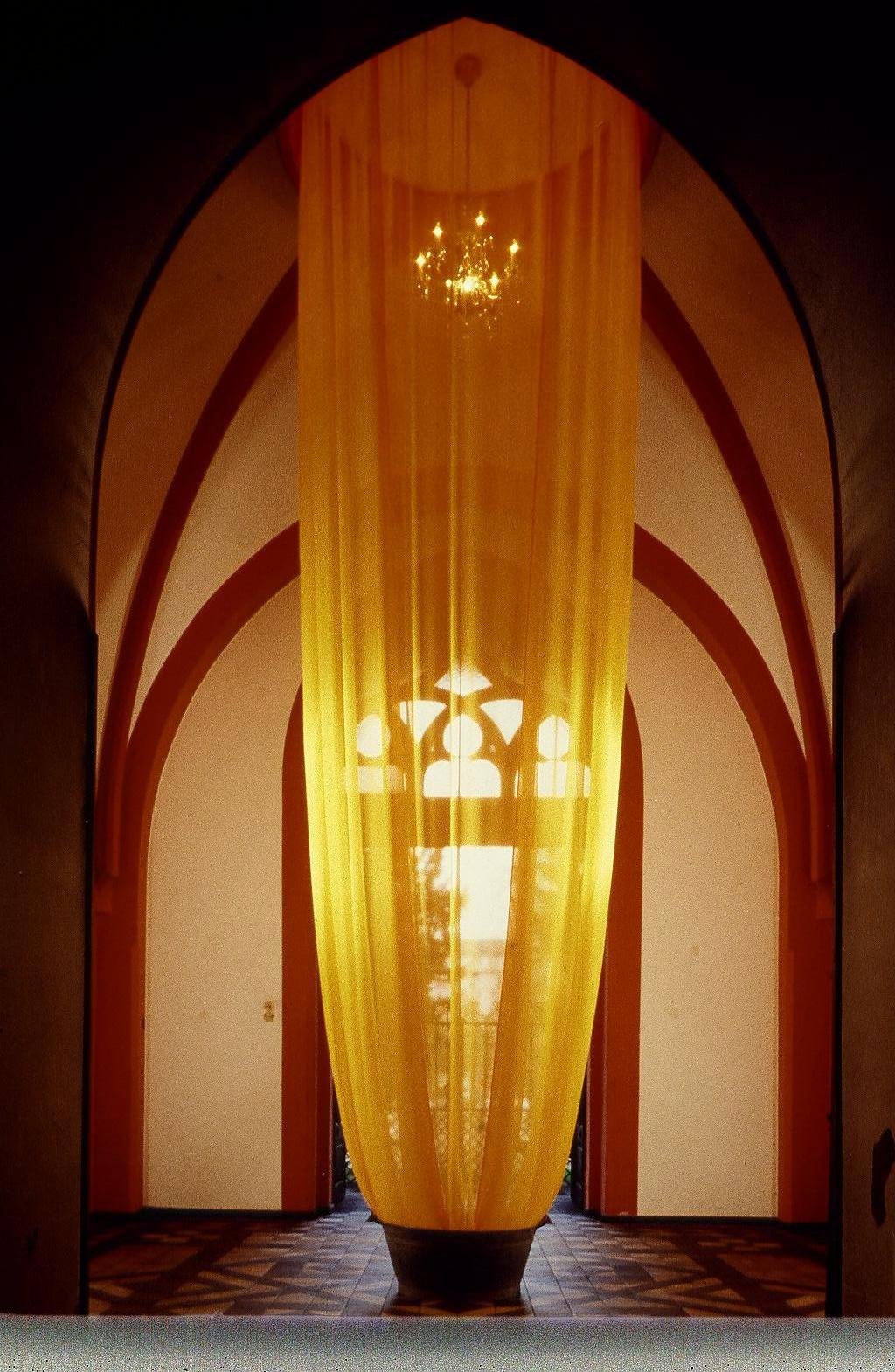 Elegant interior with arched doorways, a tall yellow curtain or fabric sculpture in the center, a chandelier hanging from the ceiling, and sunlight streaming through a window.