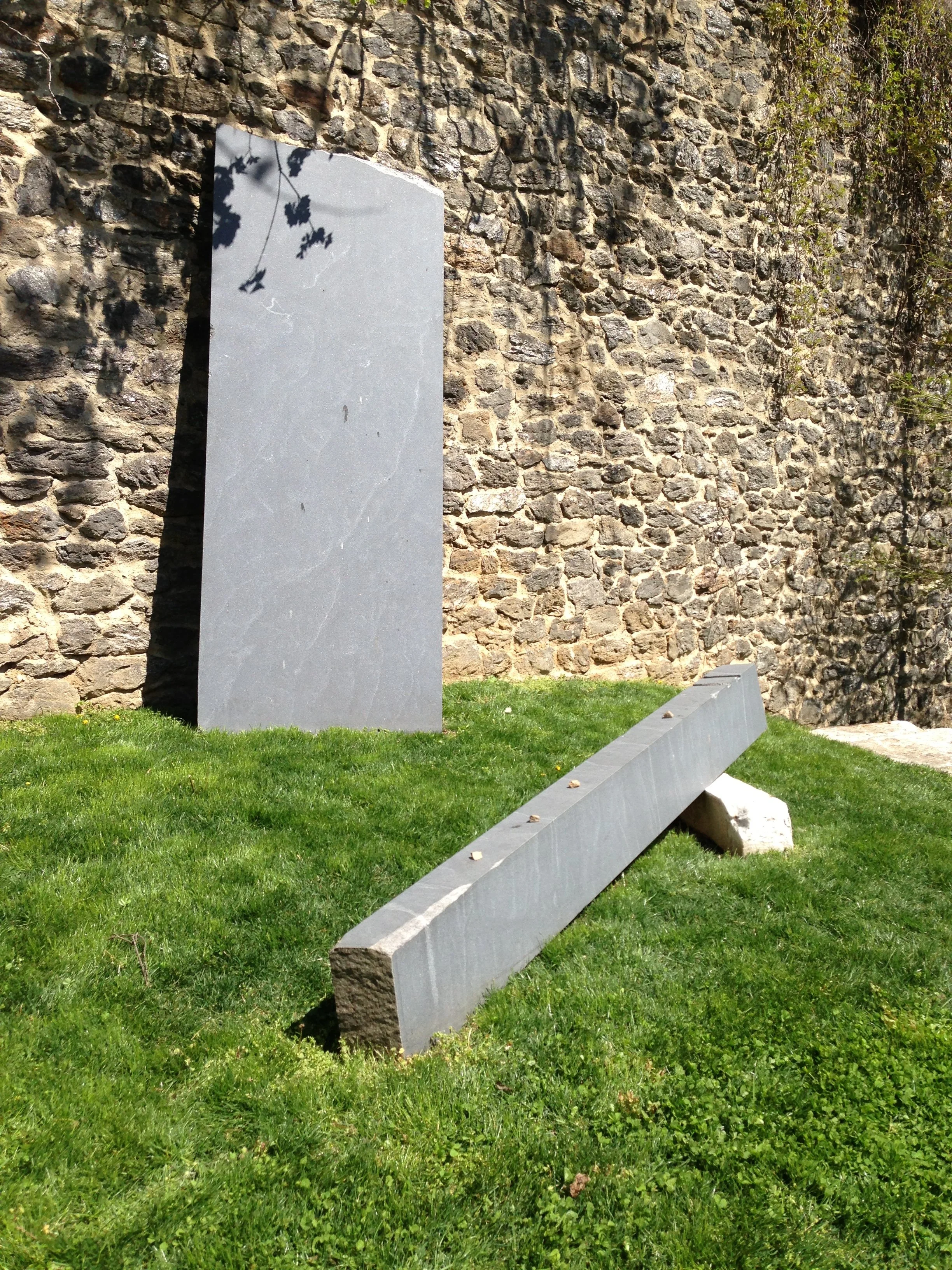 A stone wall with shadows of tree branches, modern gray stone monuments, and green grass in the foreground.
