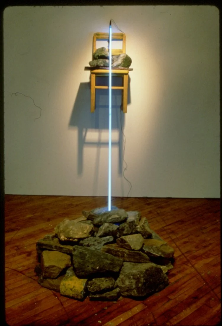 An art installation featuring a pile of rocks on the floor, with a vertical light beam extending upward through the rocks to a floating wooden chair mounted on the wall. The chair has a pillow and some rocks on it, and there is an electric cord hangi