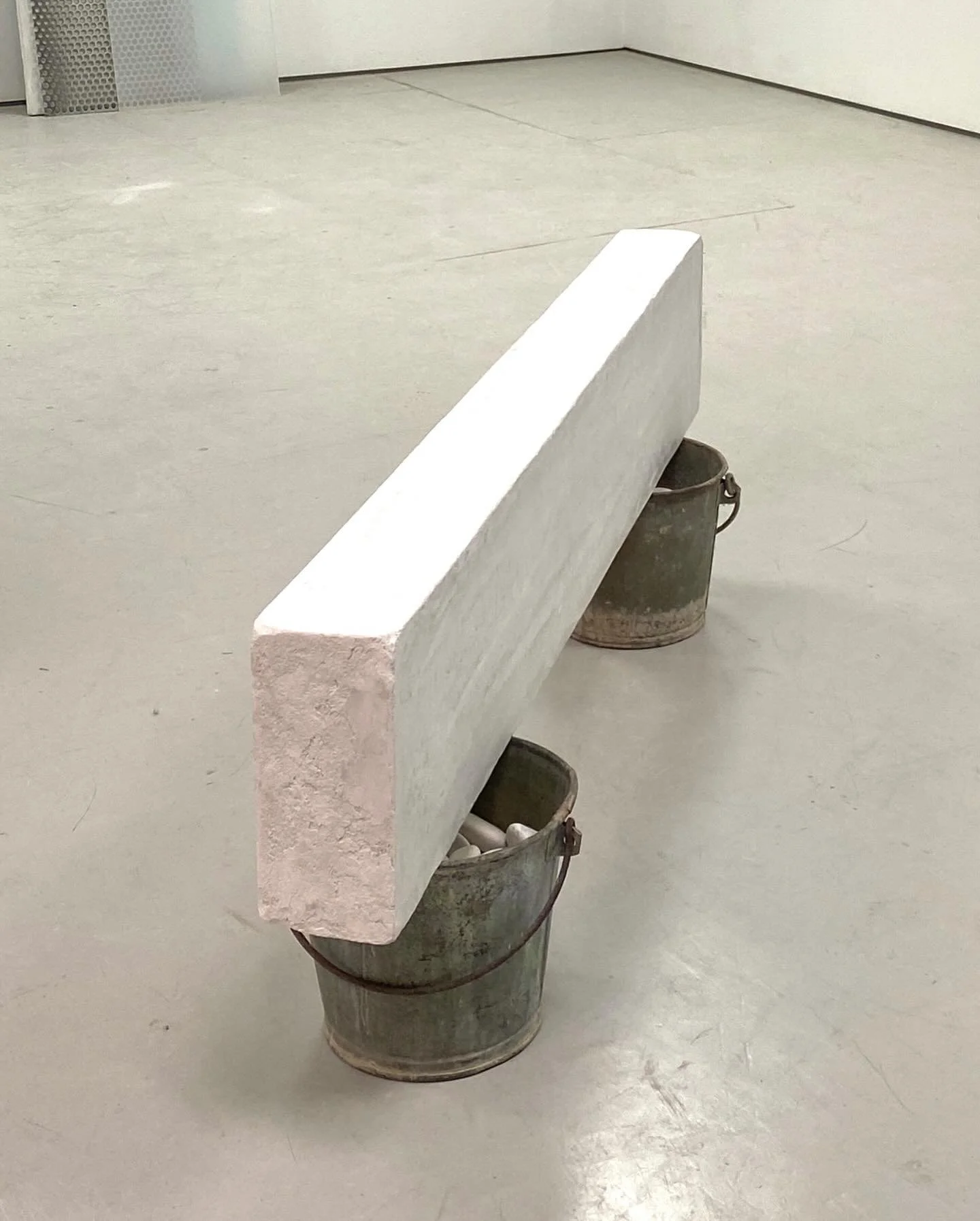 A large rectangular white stone slab resting on two metal buckets, one on each side, with the buckets filled with white objects, in an empty room with plain light-colored walls and floor.