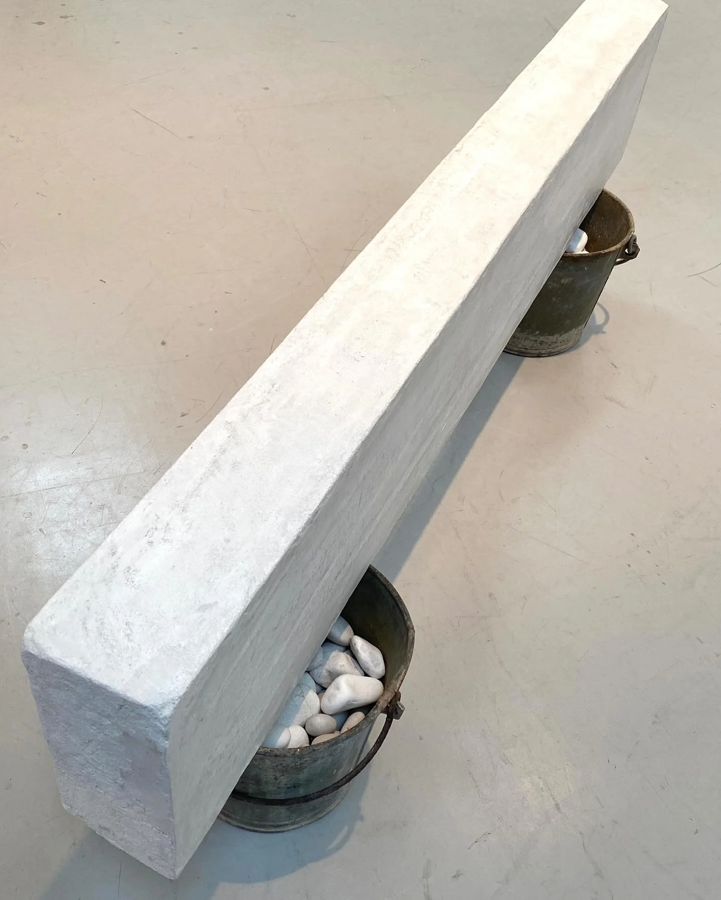 A long rectangular white stone slab is balanced over two metal buckets filled with white pebbles, on a concrete floor.