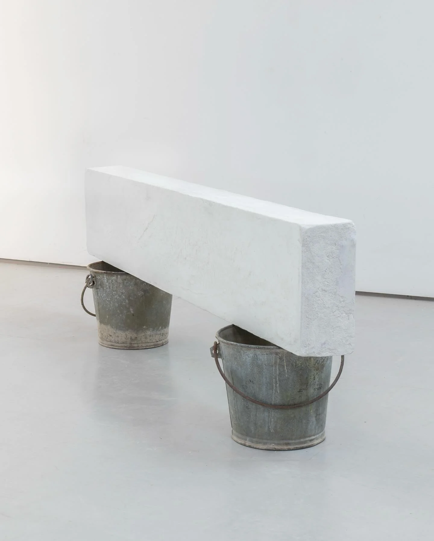 A white rectangular sculpture balanced on two metal buckets in a minimalistic white gallery space.