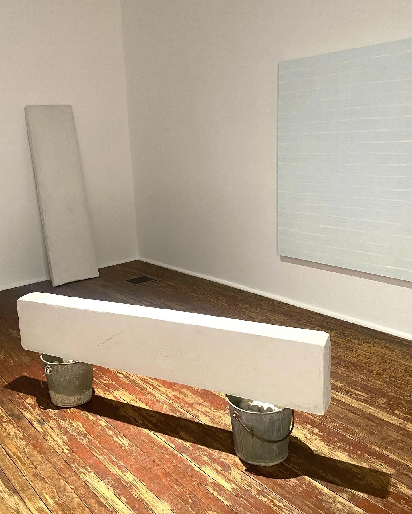 An art installation features a long, white rectangular object resting on two metal buckets filled with rocks, situated on a wooden floor in a gallery space with white walls and a large light-colored artwork hanging on the right wall.