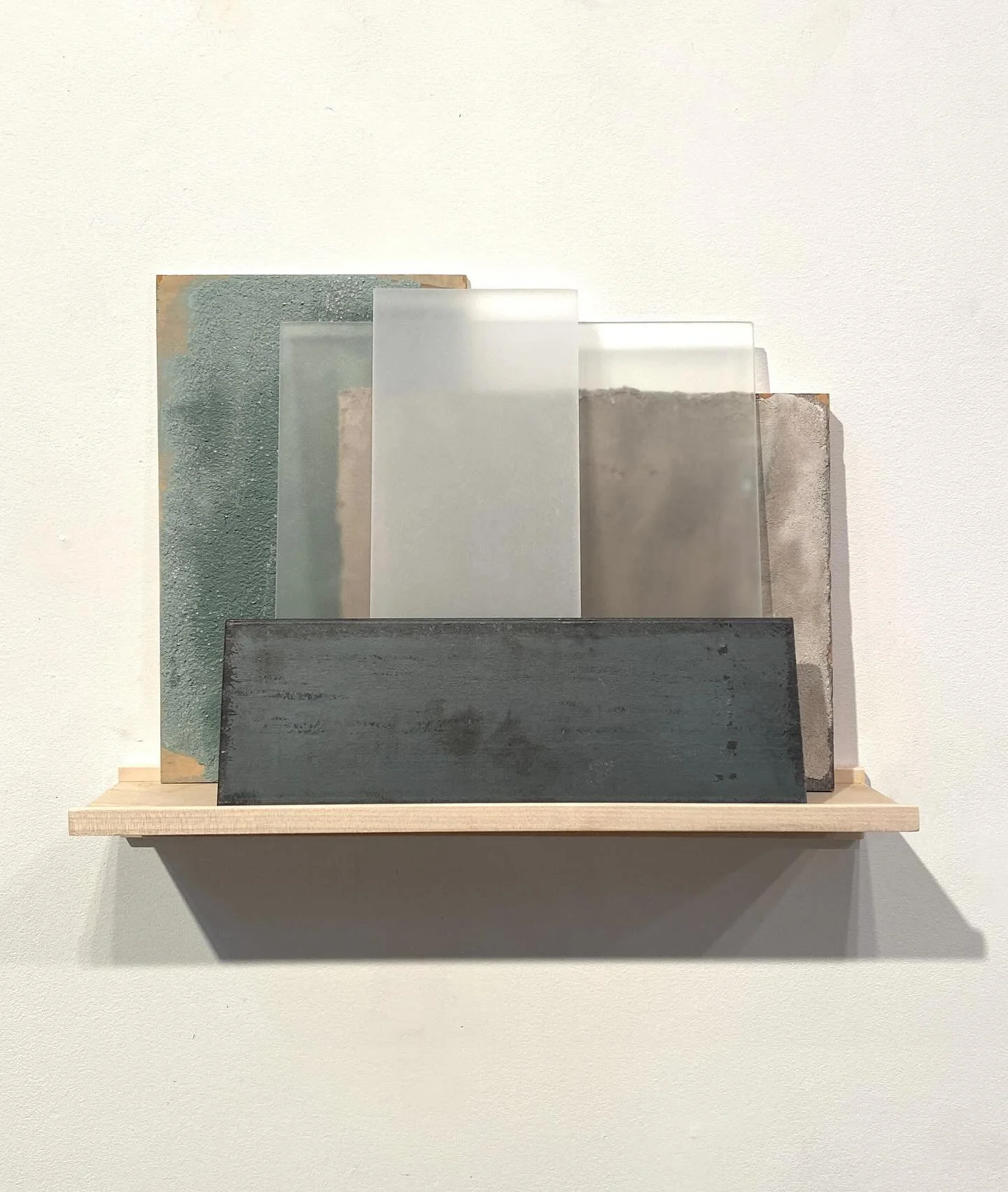 A display of six rectangular tiles with different textures and colors, arranged on a wooden shelf against a white wall.