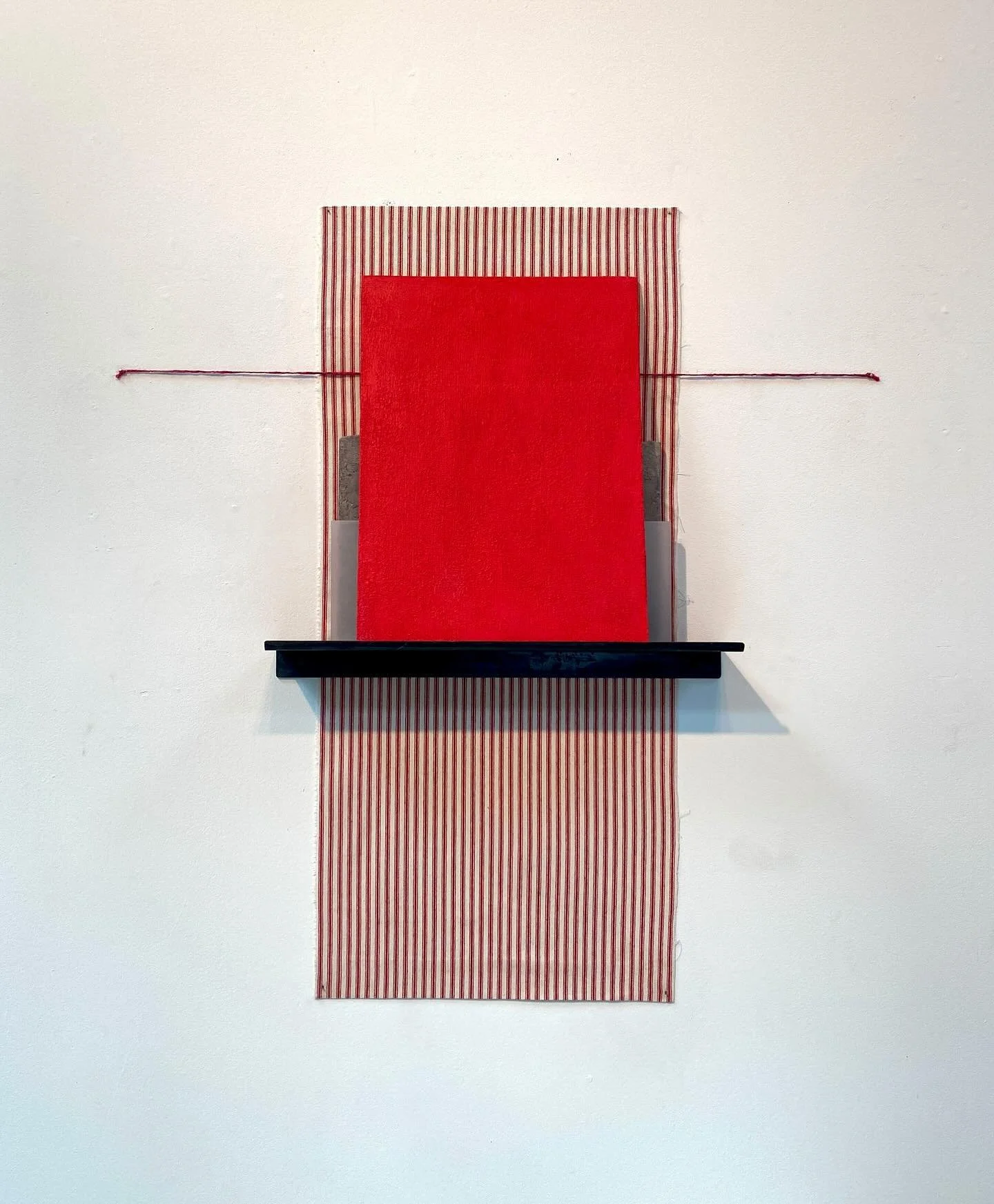 An art installation on a white wall featuring vertically striped red and white fabric extending downward, with a horizontal black shelf in front of a layered background of red and patterned fabric, and thin red string stretched horizontally across th