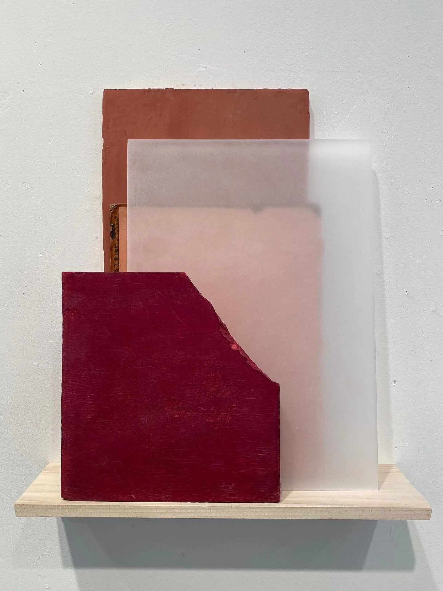 Abstract artwork consisting of three overlapping rectangular panels in shades of red, pink, and beige, mounted on a wall with a wooden shelf below.