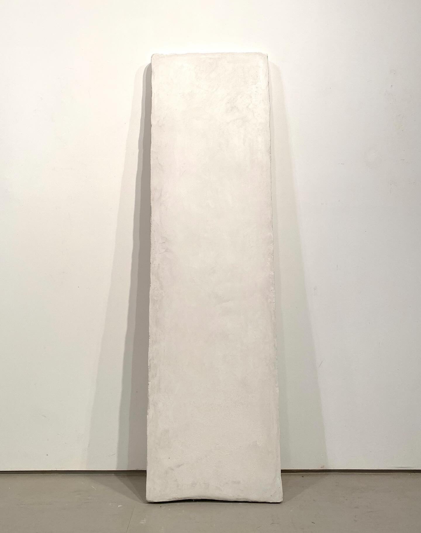 A tall white rectangular sculpture on a white base, placed against a white wall in a gallery setting.