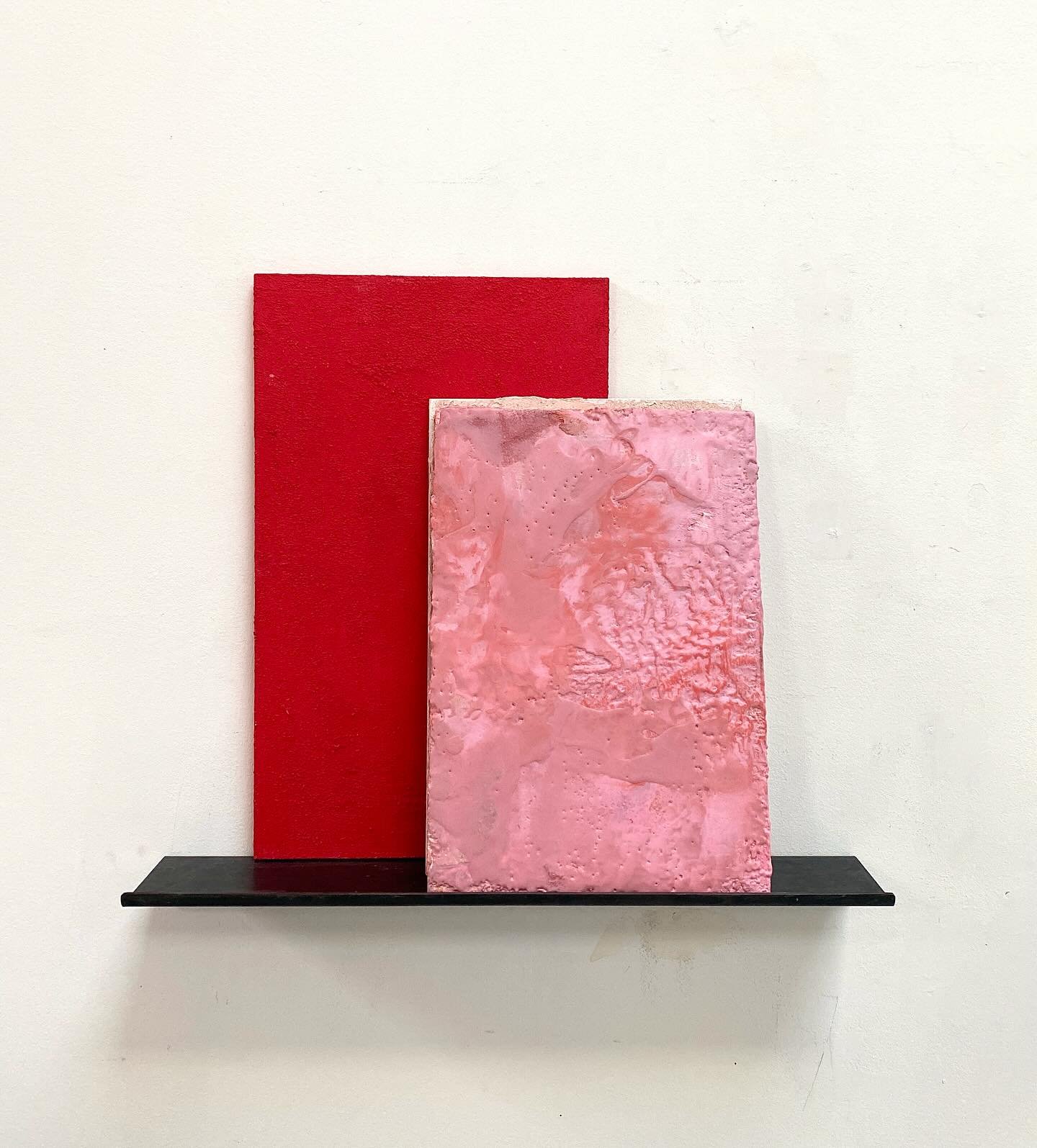 Abstract art display with a tall red rectangular panel behind a shorter pink textured rectangular panel, mounted on a black shelf against a plain white wall.
