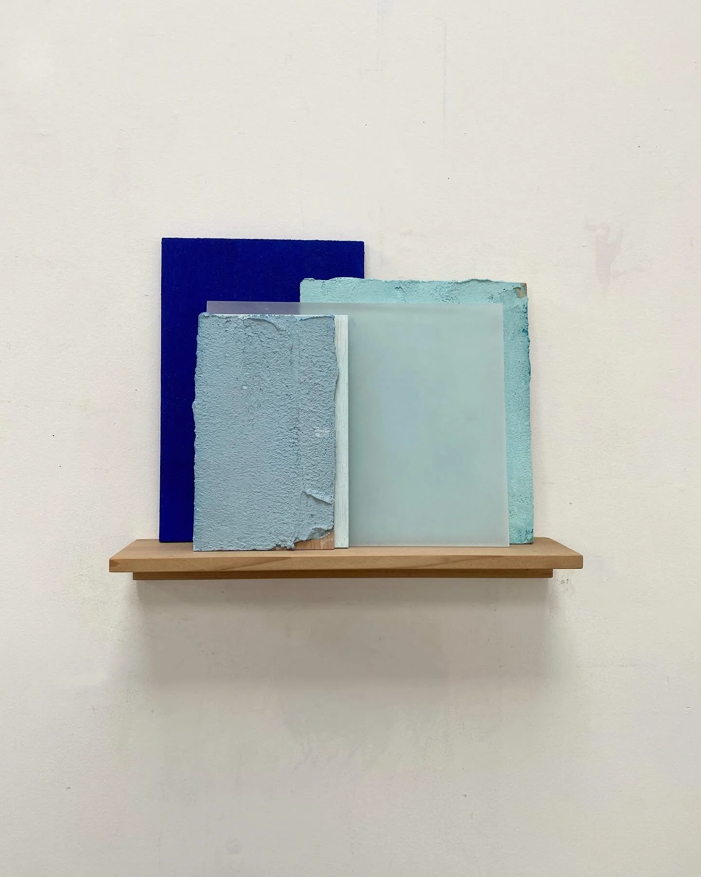 Four different textured and colored glass or acrylic panels leaning against a white wall on a wooden shelf.