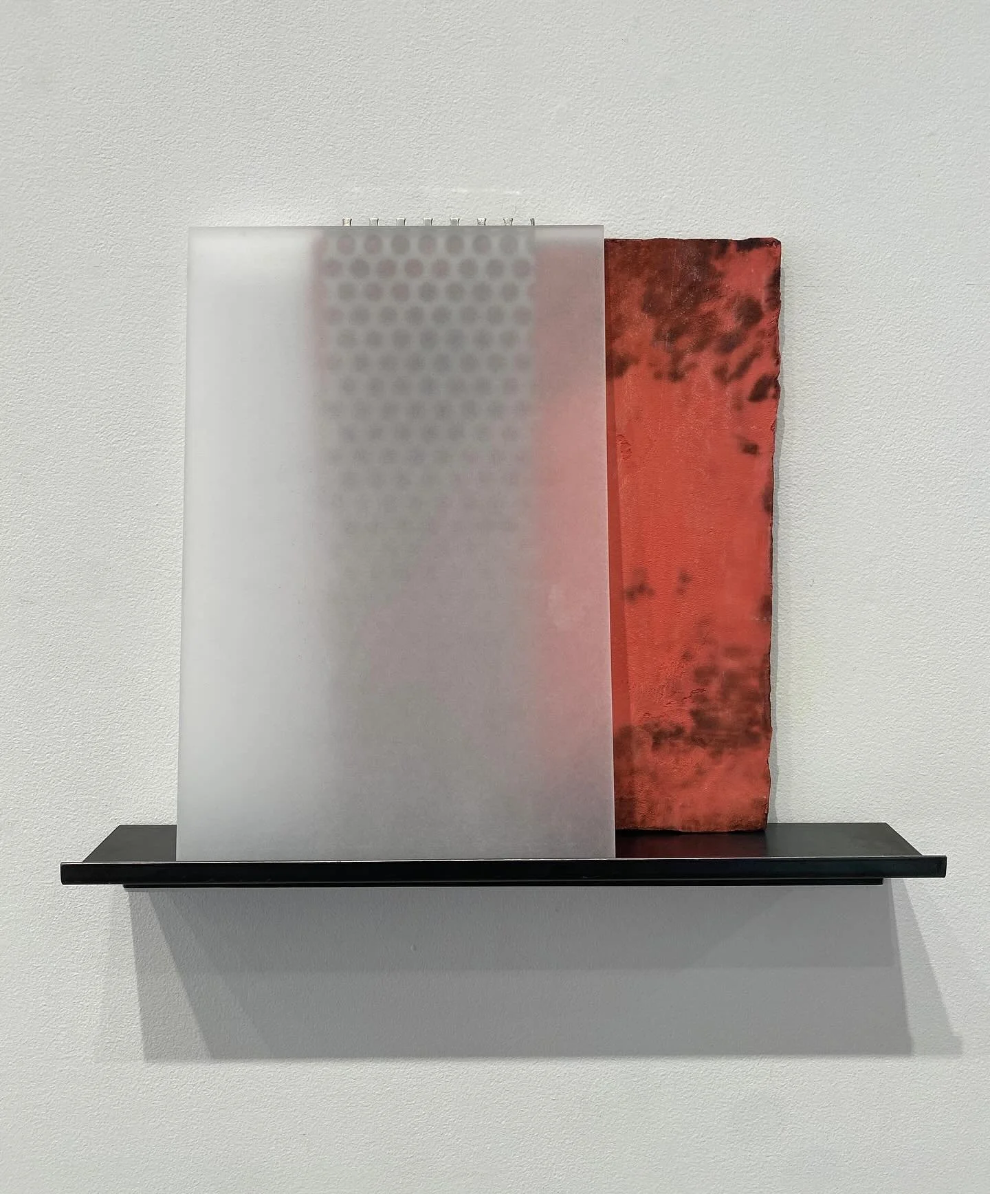Mixed media artwork featuring a semi-transparent white panel with a pattern of small circular holes on the left and a weathered red metal panel on the right, mounted on a black shelf.