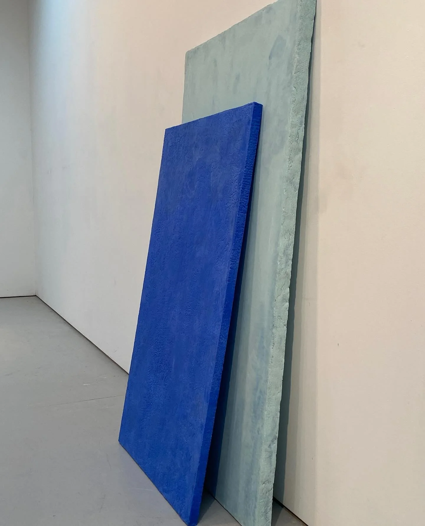 Two large rectangular panels, one painted bright blue and the other in a textured light gray, leaning against white gallery walls in an art installation or museum setting.