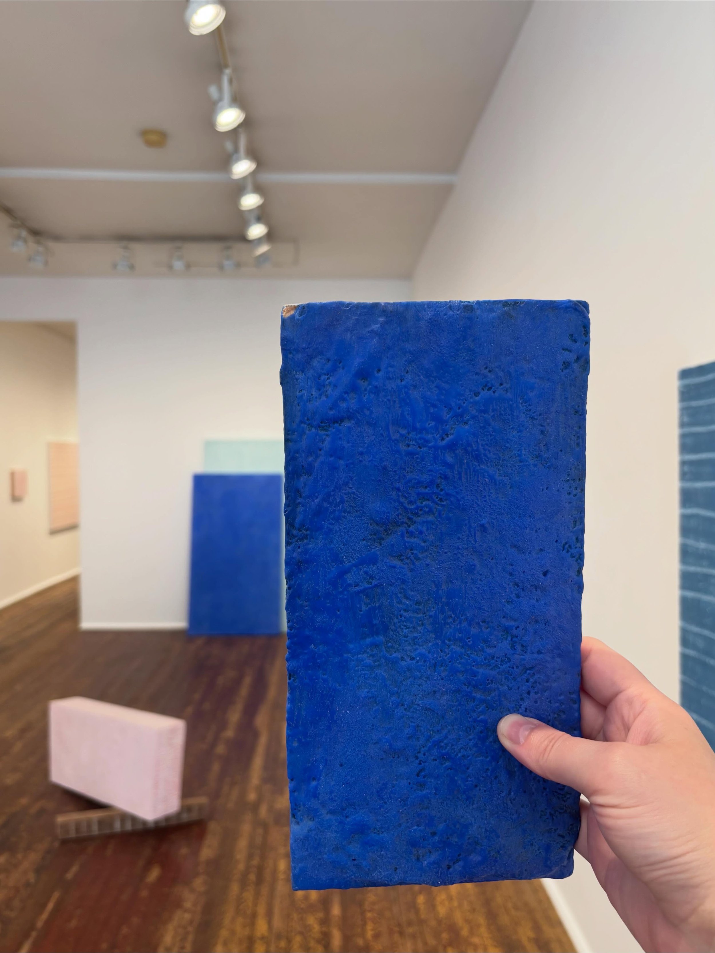 A person holding a large blue rectangular object in an art gallery. The background shows white walls with abstract artwork and track lighting on the ceiling.
