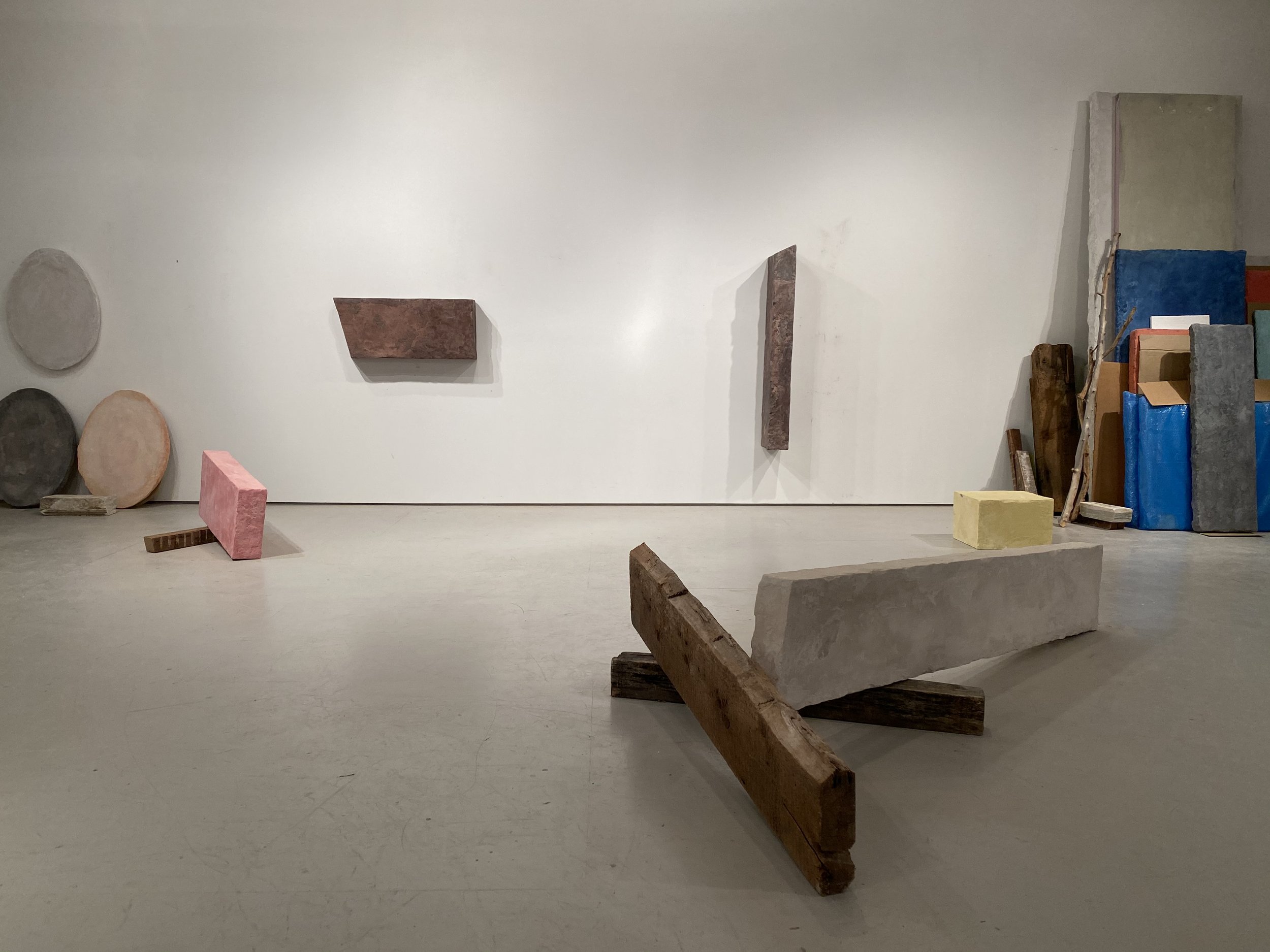 Contemporary art installation with variously shaped and colored stone, wood, and concrete blocks on a white gallery wall and floor.