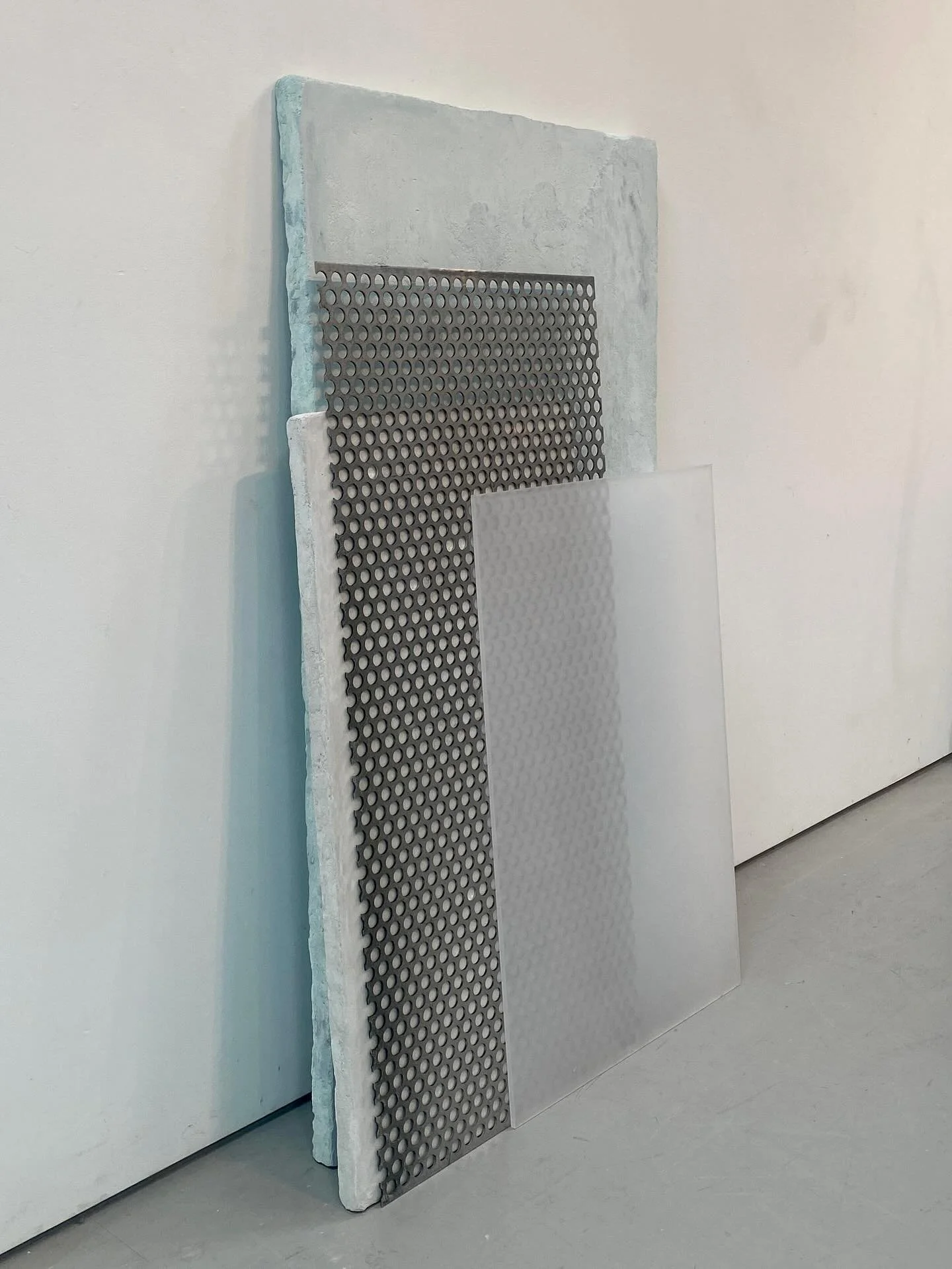 Three large panels leaning against a white wall. The first panel is made of foam with a rough texture. The second panel has a grid of circular holes. The third panel is a translucent panel with a smaller circular pattern.