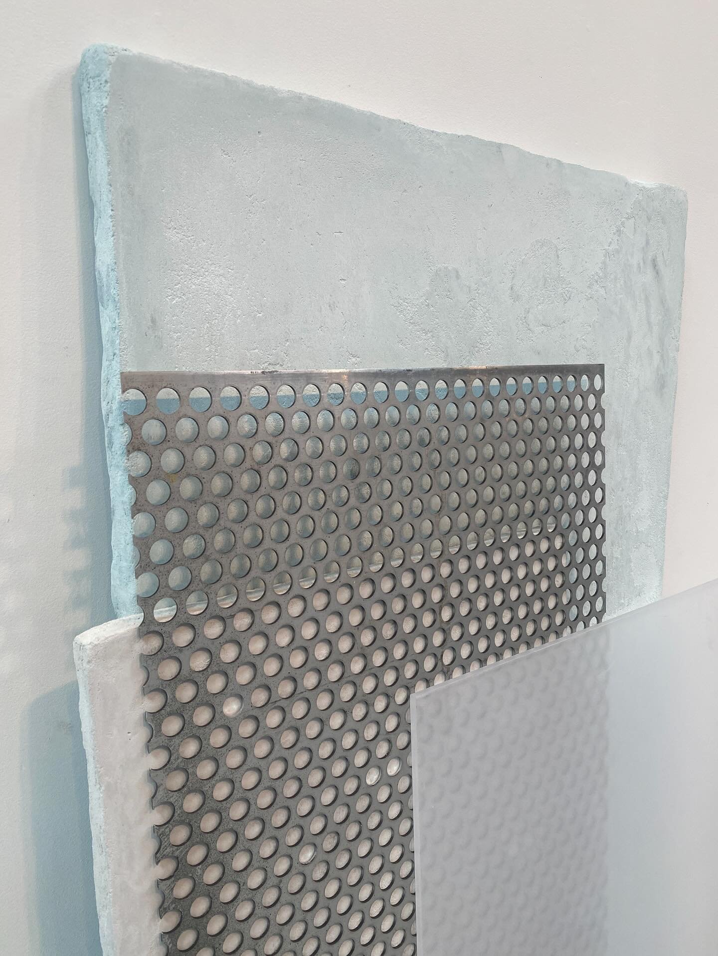 A metal grate with circular holes leaning against a white wall, with a textured surface behind it and an additional rectangular piece of textured material below.