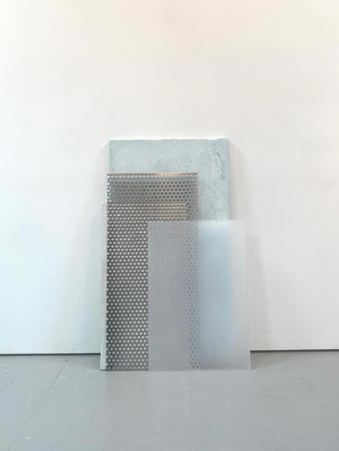 Three sheets of perforated metal and one solid metal sheet leaning against a white wall in a minimalistic space.