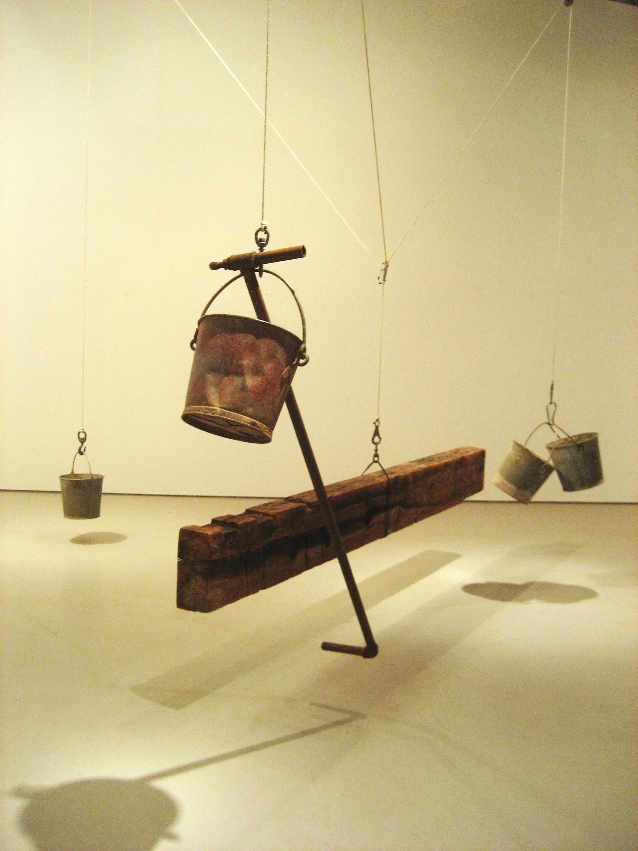 A hanging mobile sculpture featuring a small bicycle with a bucket on its handlebars, balanced on a narrow wooden plank, cast shadows on a cream-colored wall.