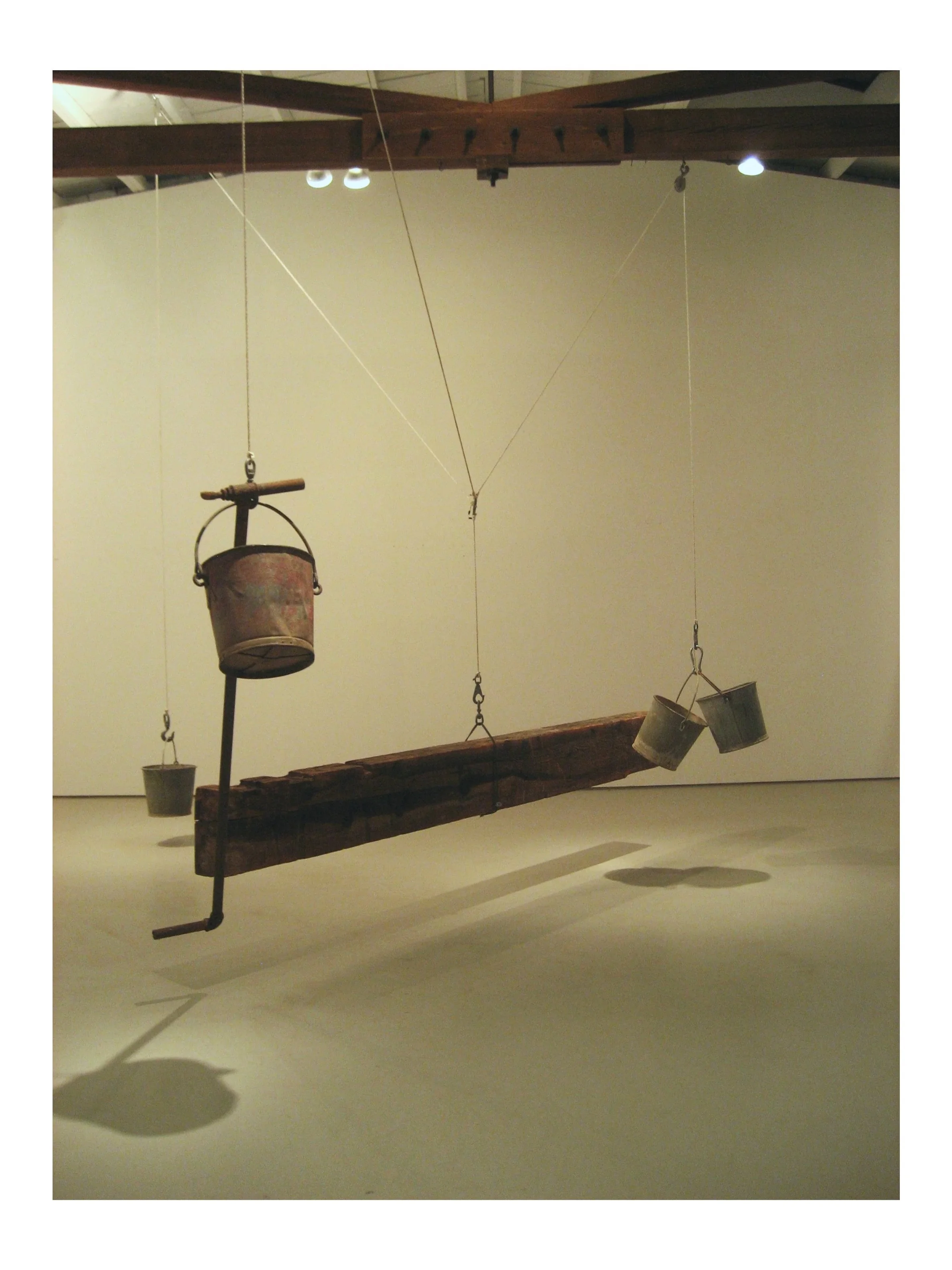 An art installation featuring a wooden plank suspended with ropes, with various hanging objects including two buckets and a metal container, all against a plain white wall.