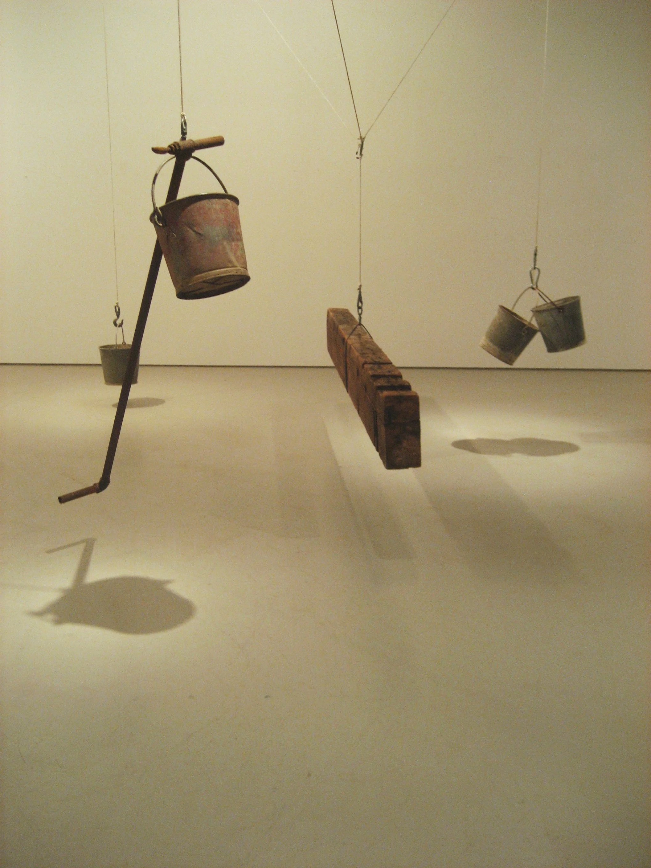 A mobile art installation consisting of three small metal buckets suspended from strings, a weathered wooden block, and a long metal rod, all hanging from the ceiling in a gallery setting.