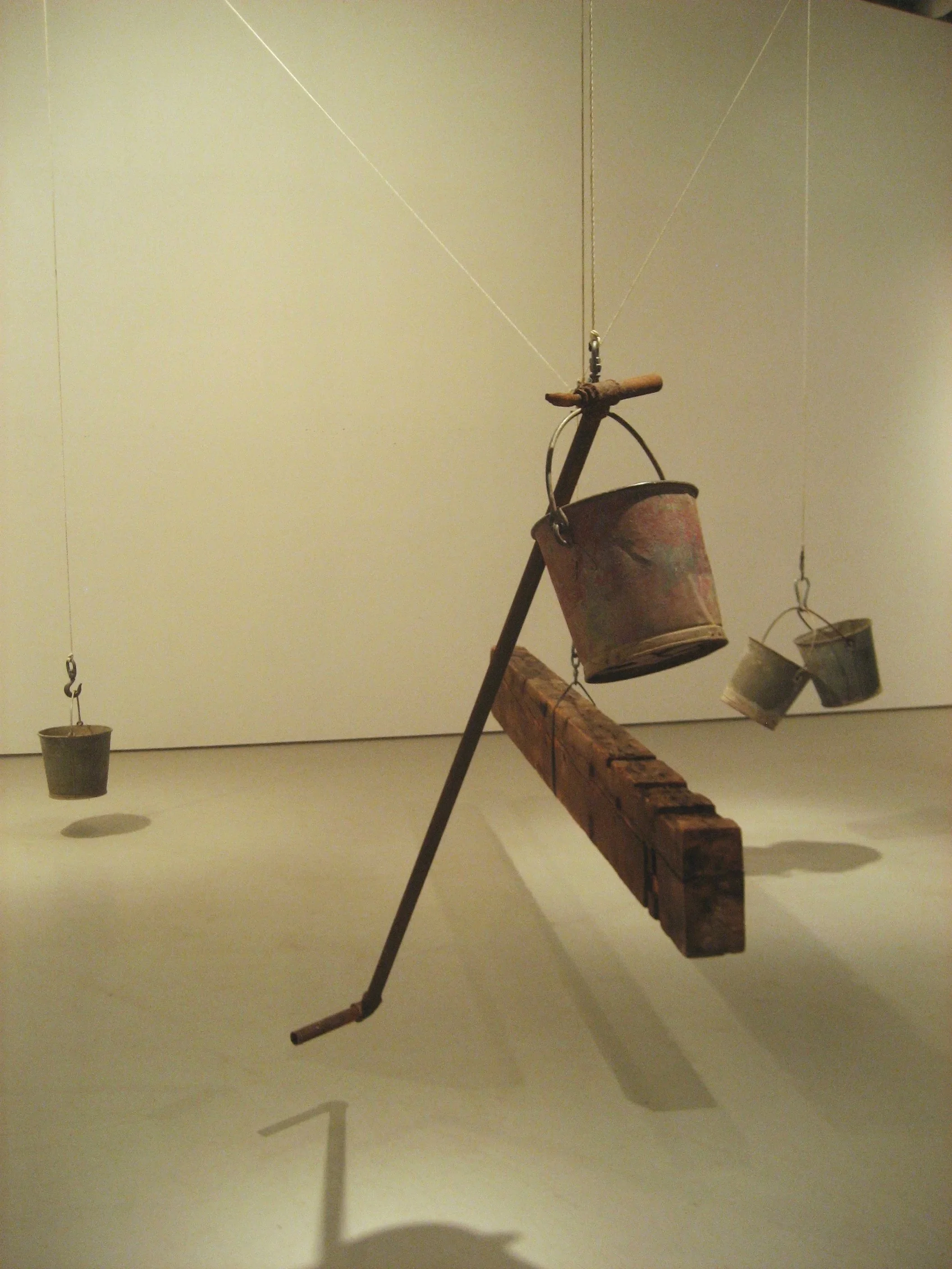 An art installation with a wooden plank suspended by strings from the ceiling, with hanging metal buckets attached to the ends of the plank and additional buckets hanging from the ceiling behind.
