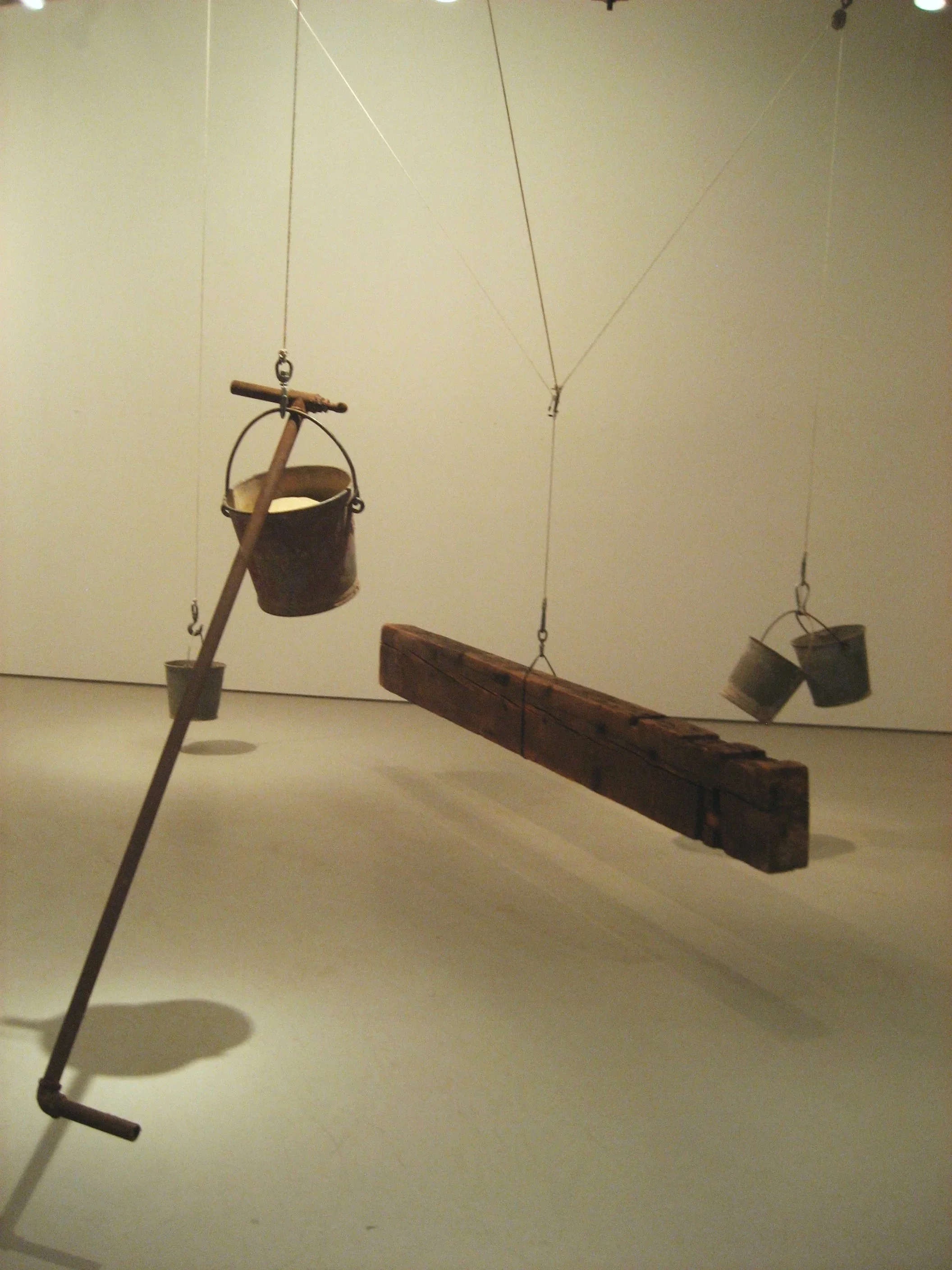An art installation with a wooden plank suspended by ropes, with small buckets hanging from the ropes around the plank, and a wooden pole with a bucket attached to the top leaning against the plank.