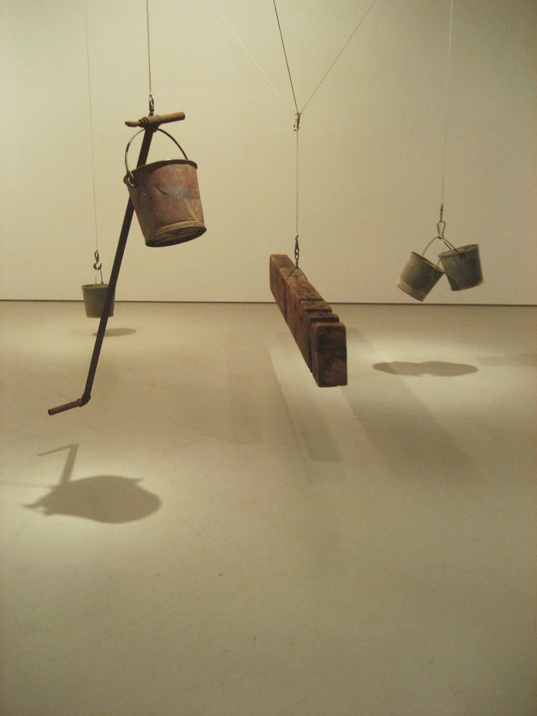 A museum installation with hanging objects, including old metal buckets, a wooden block, and a long metal rod, casting shadows on a light-colored wall and floor.