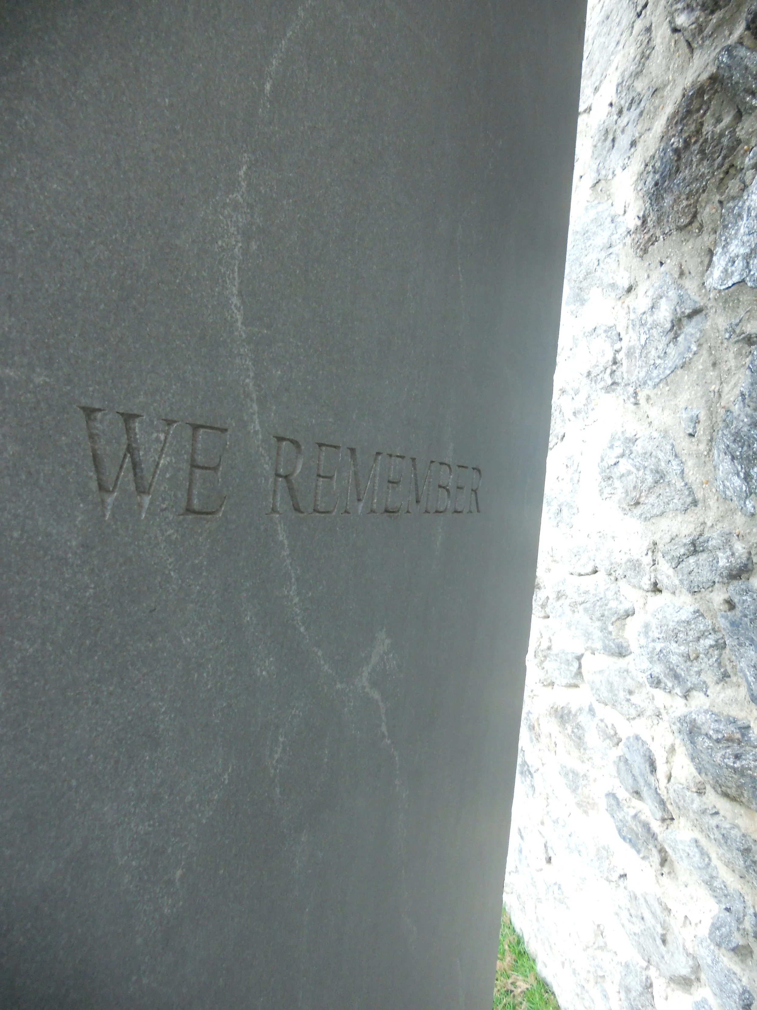 Close-up of a memorial plaque with the words "WE REMEMBER" engraved on it, placed outdoors on a grassy area with rocks.