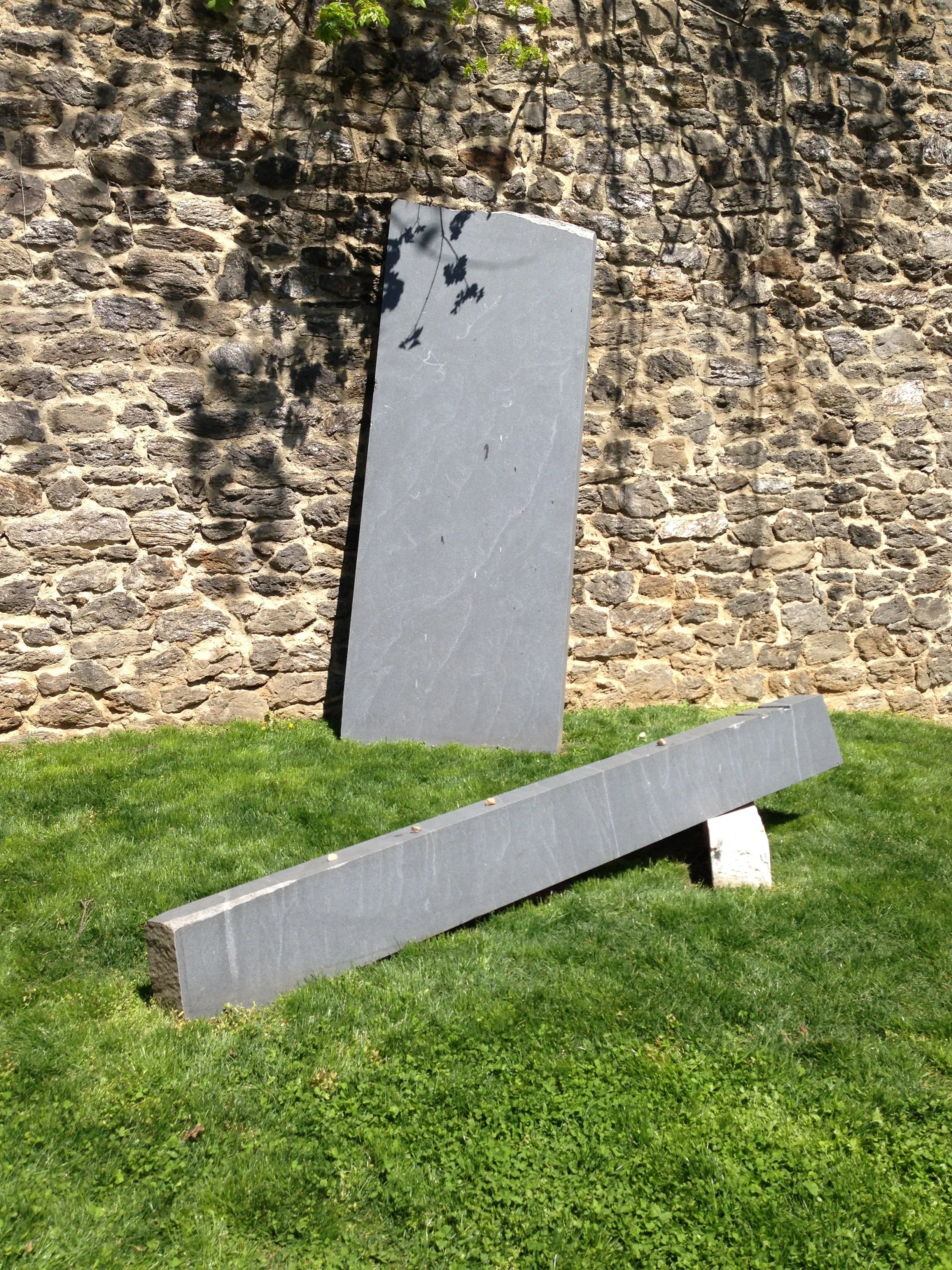 A stone wall with a shadow of a plant cast on it. In front of the wall, two large, rectangular, gray stones are positioned on a green grass lawn, with one stone leaning against the wall and the other lying flat on the ground.