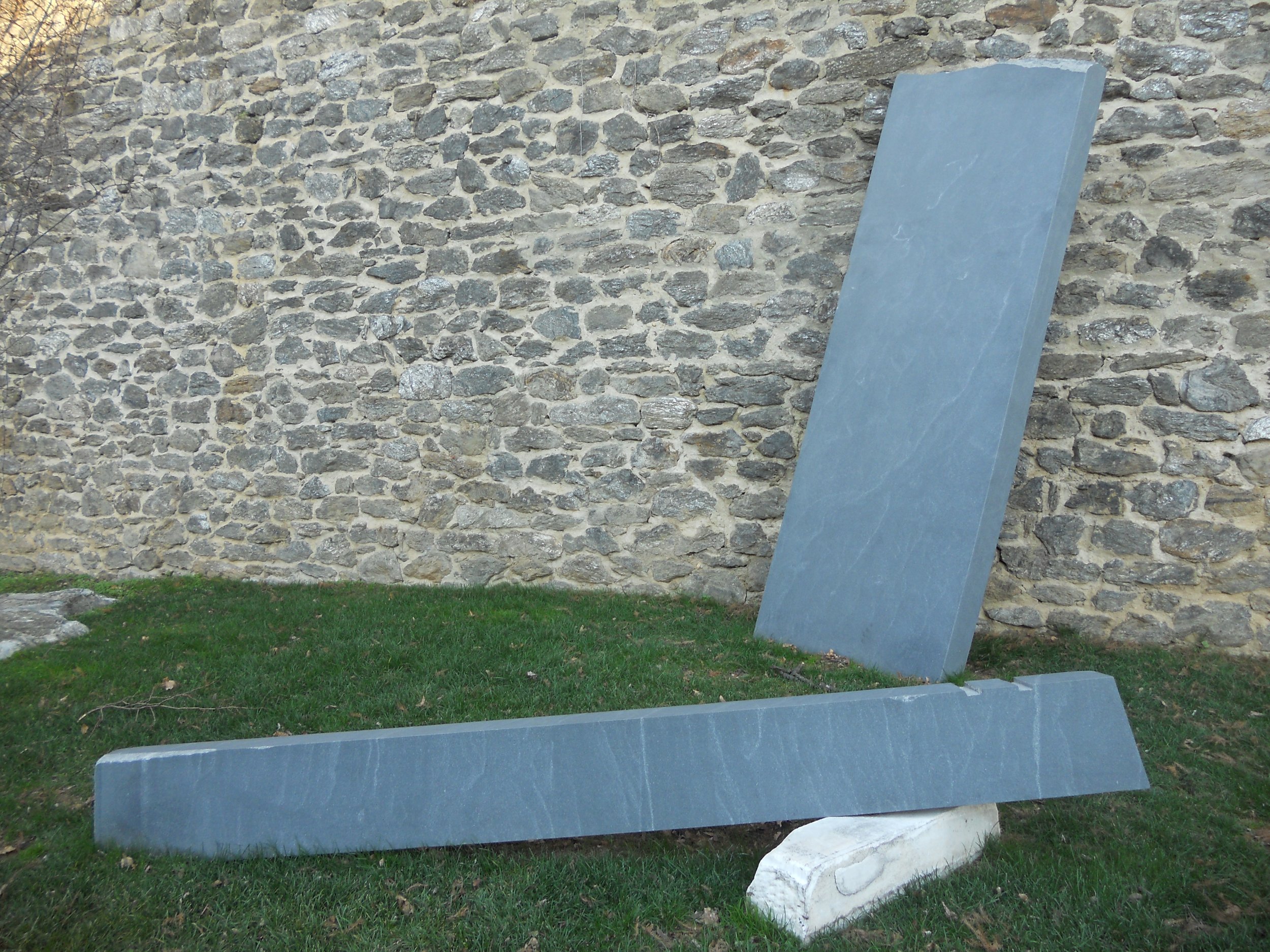 A stone wall with a blue leaning slab and a large white rock on grass in front.