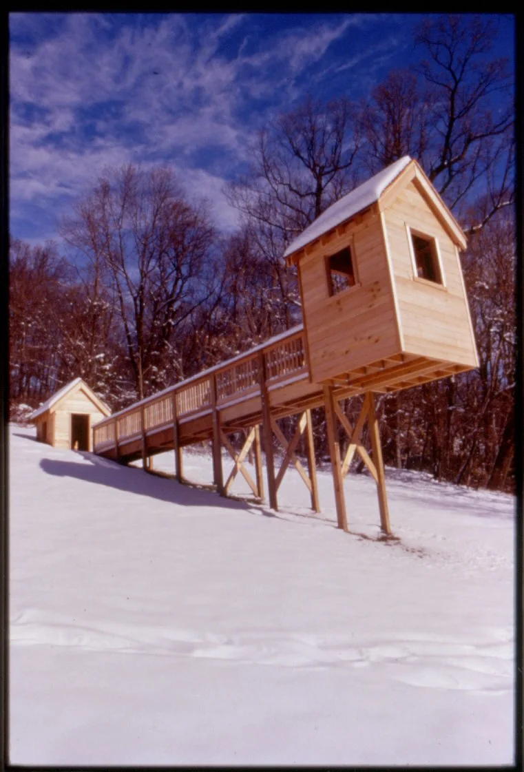 A snow-covered landscape with a wooden treehouse on stilts connected by a ramp, and a smaller wooden structure in the background, under a partly cloudy sky.