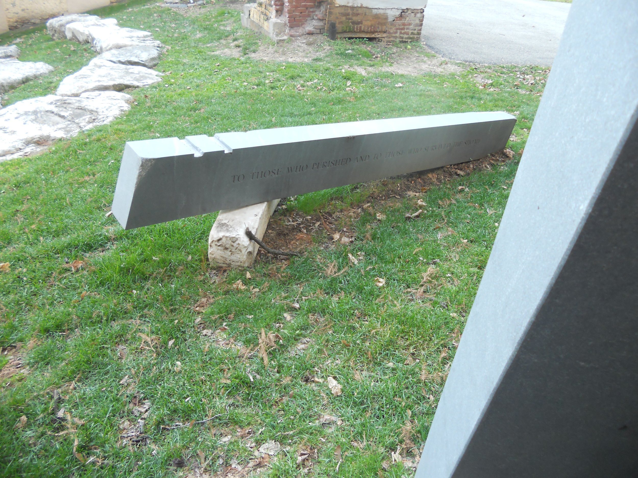 A memorial with a metal plaque mounted on a stone base, inscribed with text, placed on grass with a path and brick wall in the background.