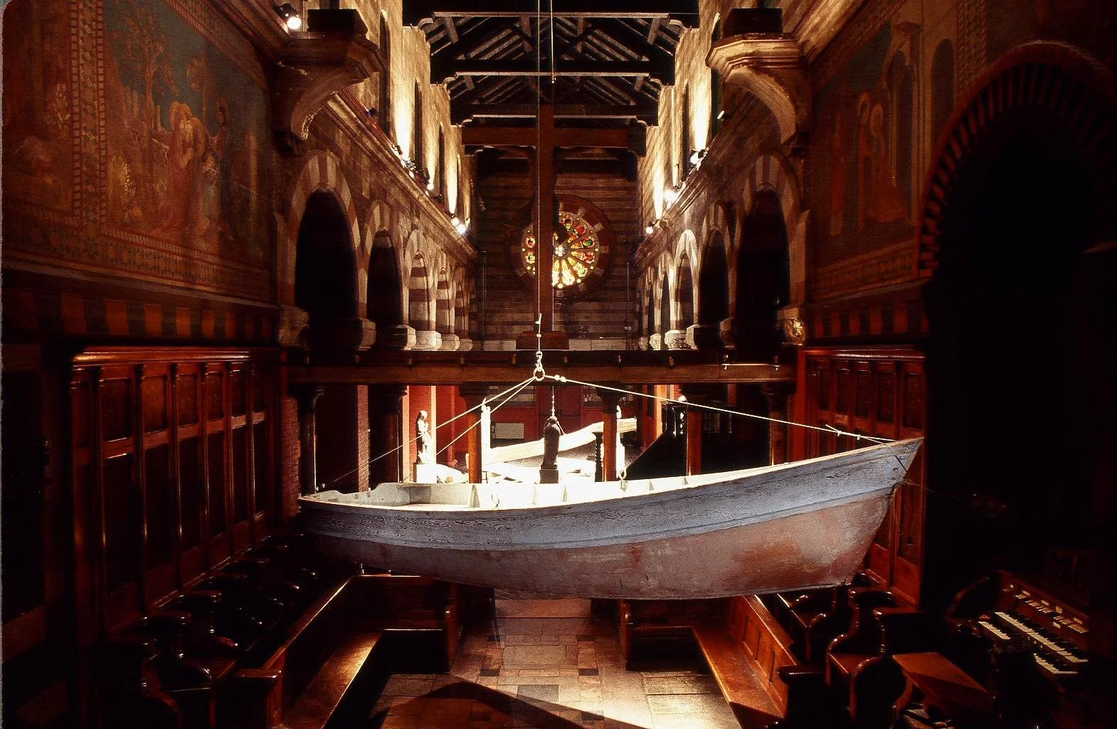 A boat hanging inside a church with wood-paneled walls and high arched ceilings. The boat is suspended by ropes, with an organ visible to the right. Light filters through a stained glass window at the back of the church.