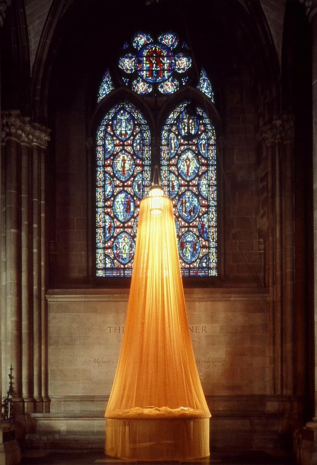 A large stained glass church window with intricate blue, red, and yellow designs, illuminated by a bright yellow fabric-covered light hanging in front of it.