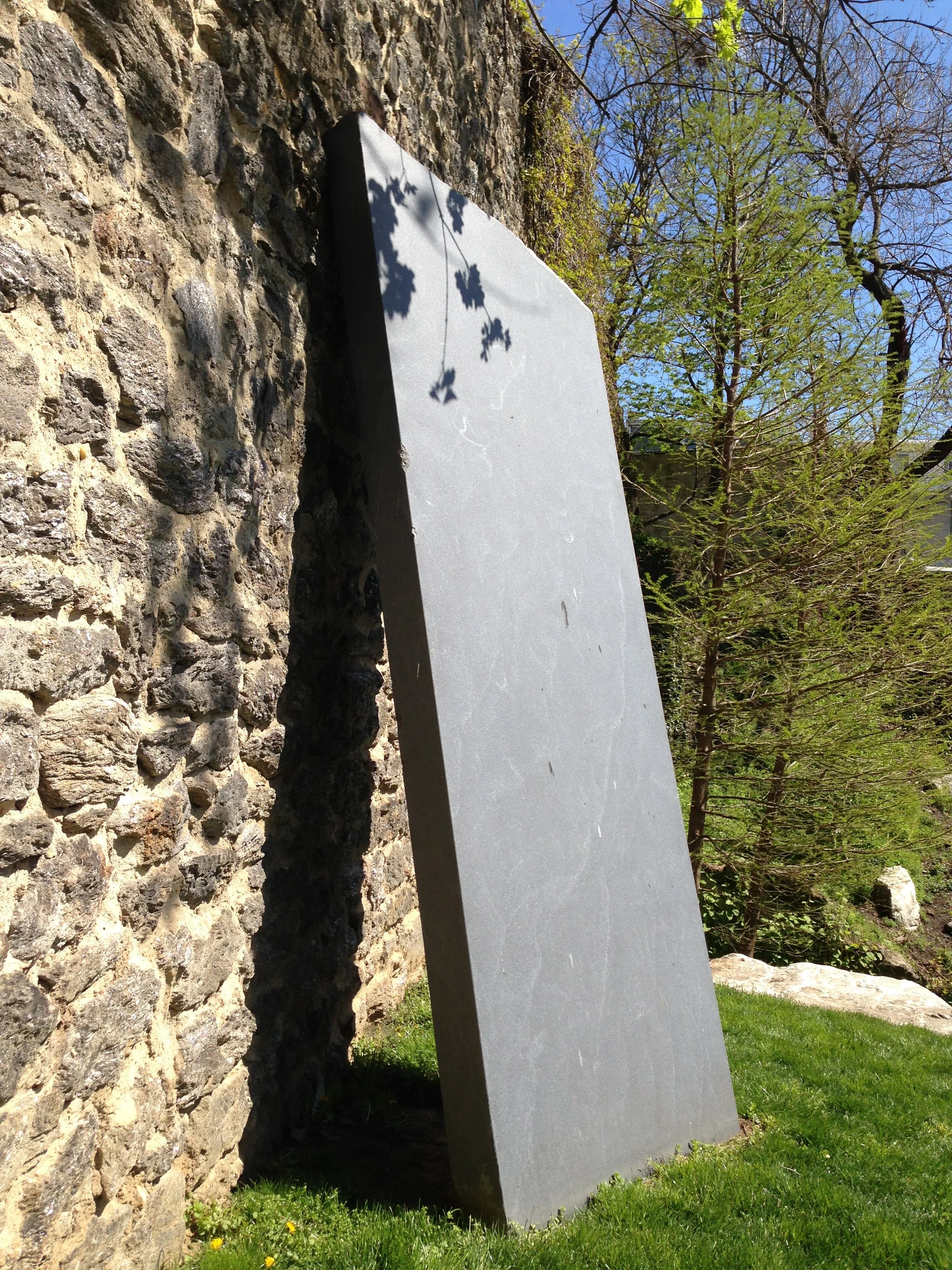 A large, rectangular, gray stone or concrete monument leaning against a stone wall in an outdoor setting, with green grass and trees in the background under a clear blue sky.