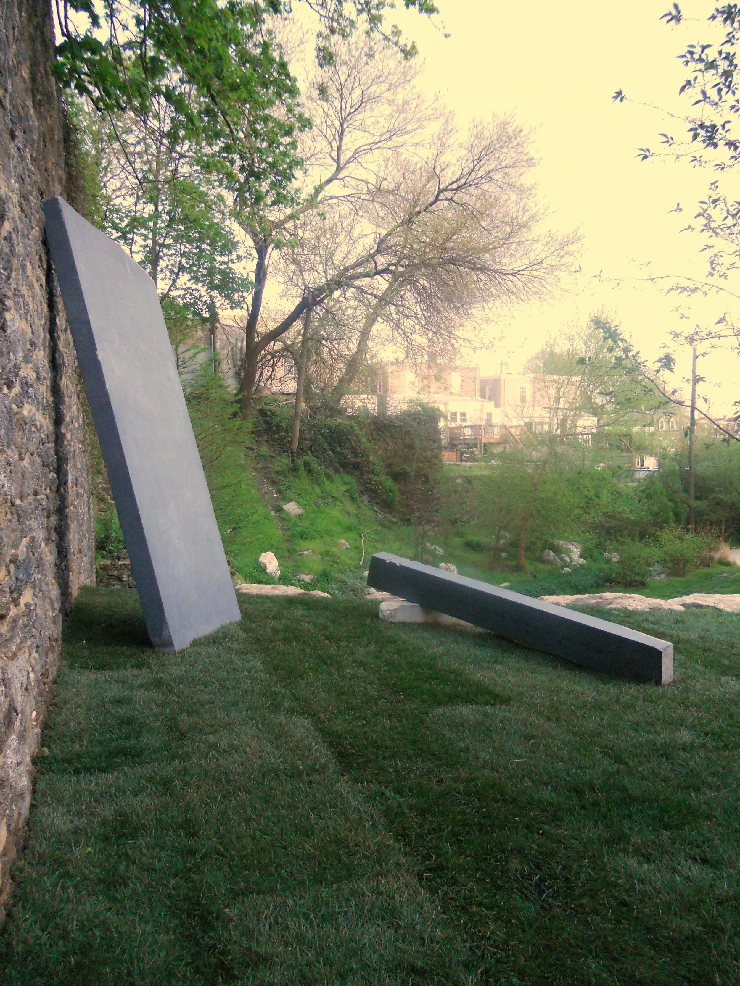 Large gray rectangular stone leaning against a rough stone wall, with two black rectangular stones lying on a well-maintained grassy area nearby, trees with budding leaves and some leafless trees, and residential buildings visible in the background u