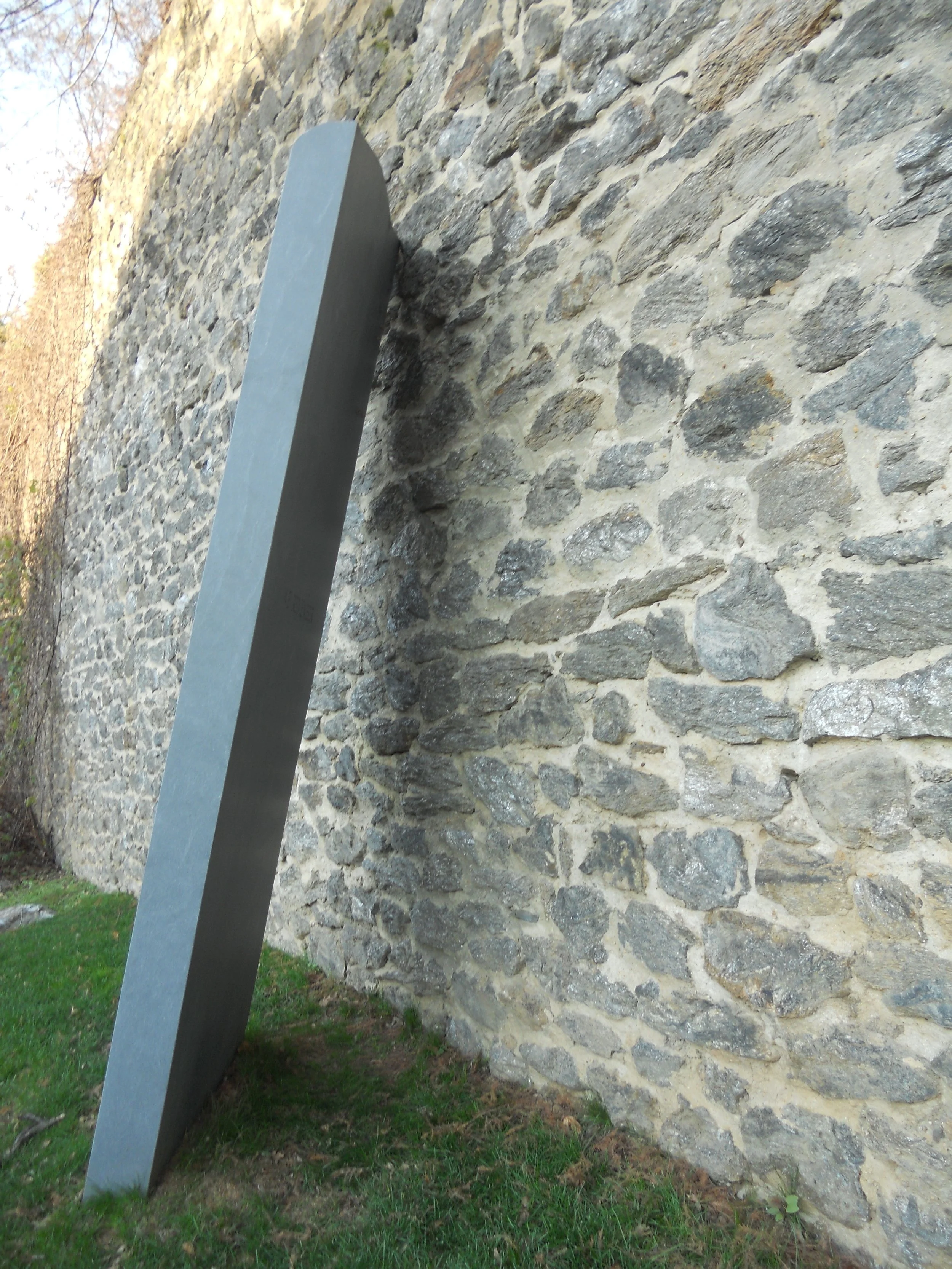 A modern gray metal sculpture leaning against a stone wall on a grassy area outdoors.