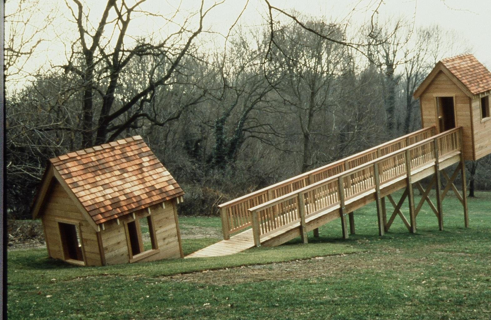 Two small wooden playhouses connected by a ramp in a grassy yard, with leafless trees in the background.