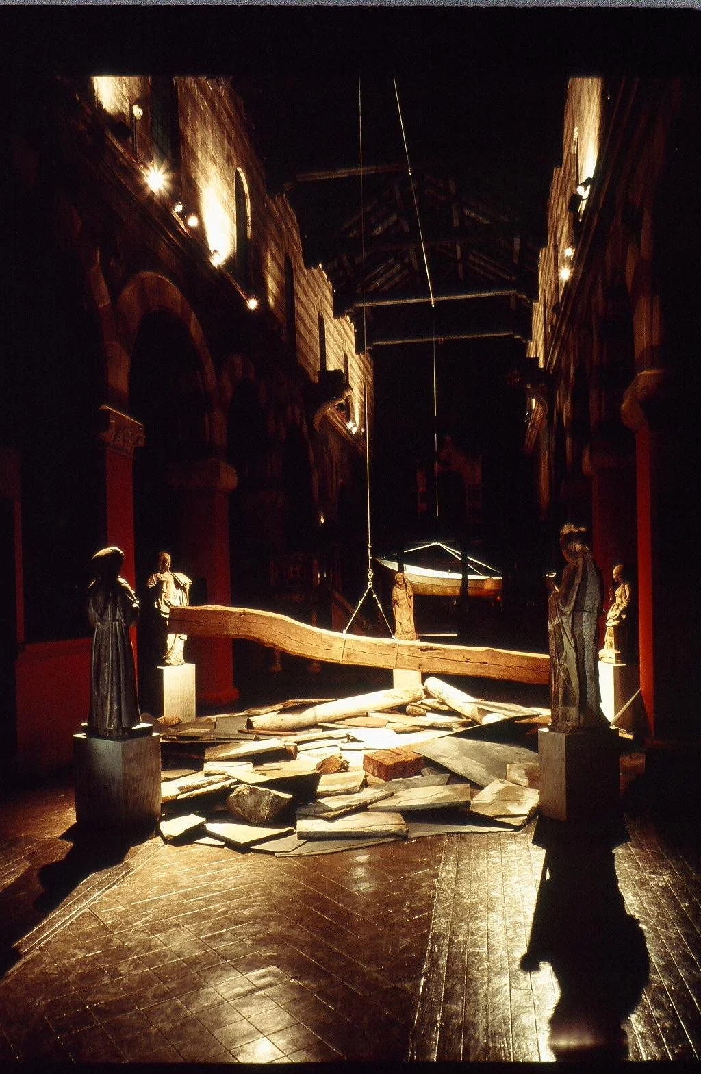 A dark, dramatic interior with a suspended wooden bridge arching over scattered debris, surrounded by statues of religious figures in a dimly lit space.