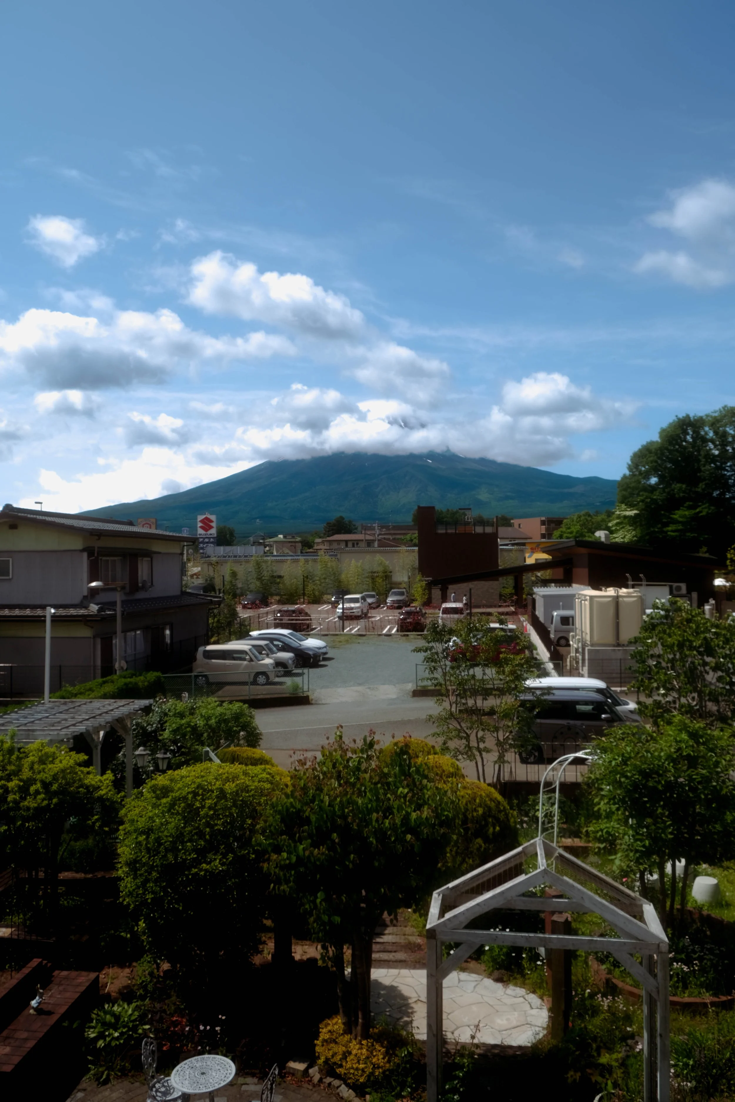 A scenic view of a mountain with a partly cloudy sky, seen from an urban area with trees, buildings, parked cars, and outdoor furniture.