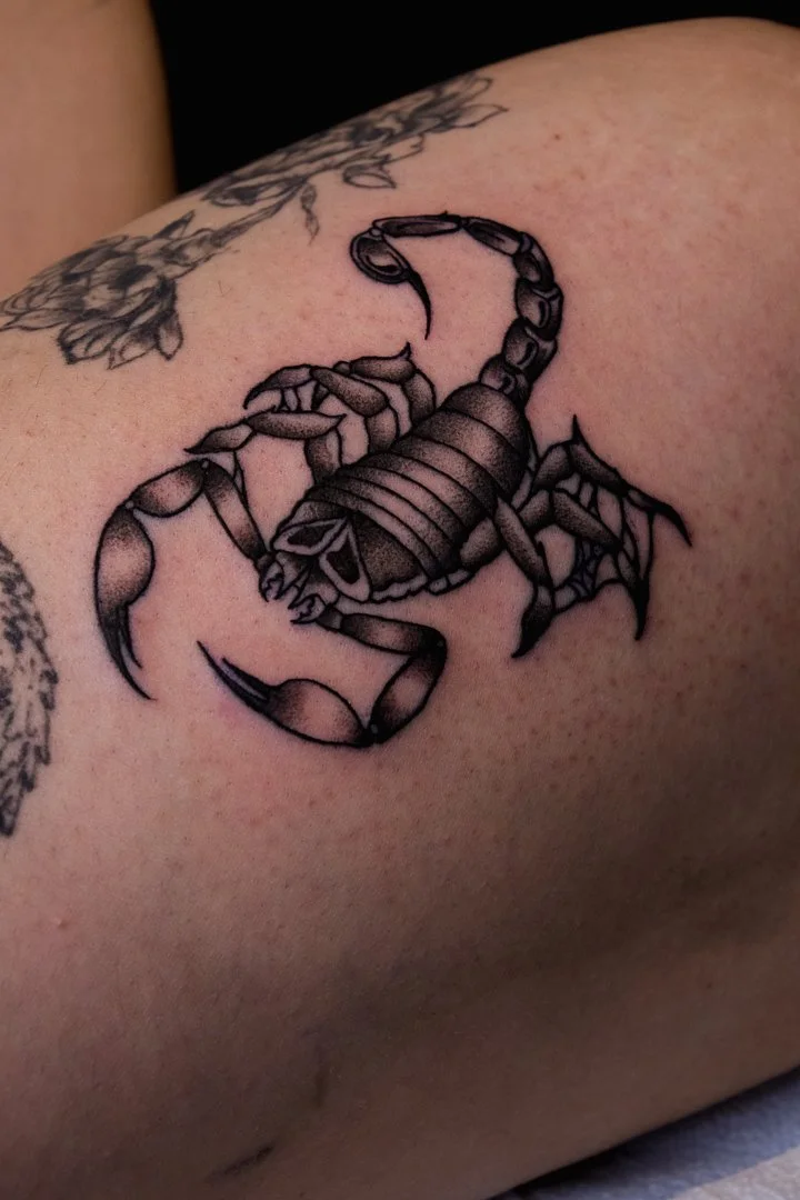 Tattoo of a black and gray scorpion on skin, with detailed claws, legs, tail, and body segments.