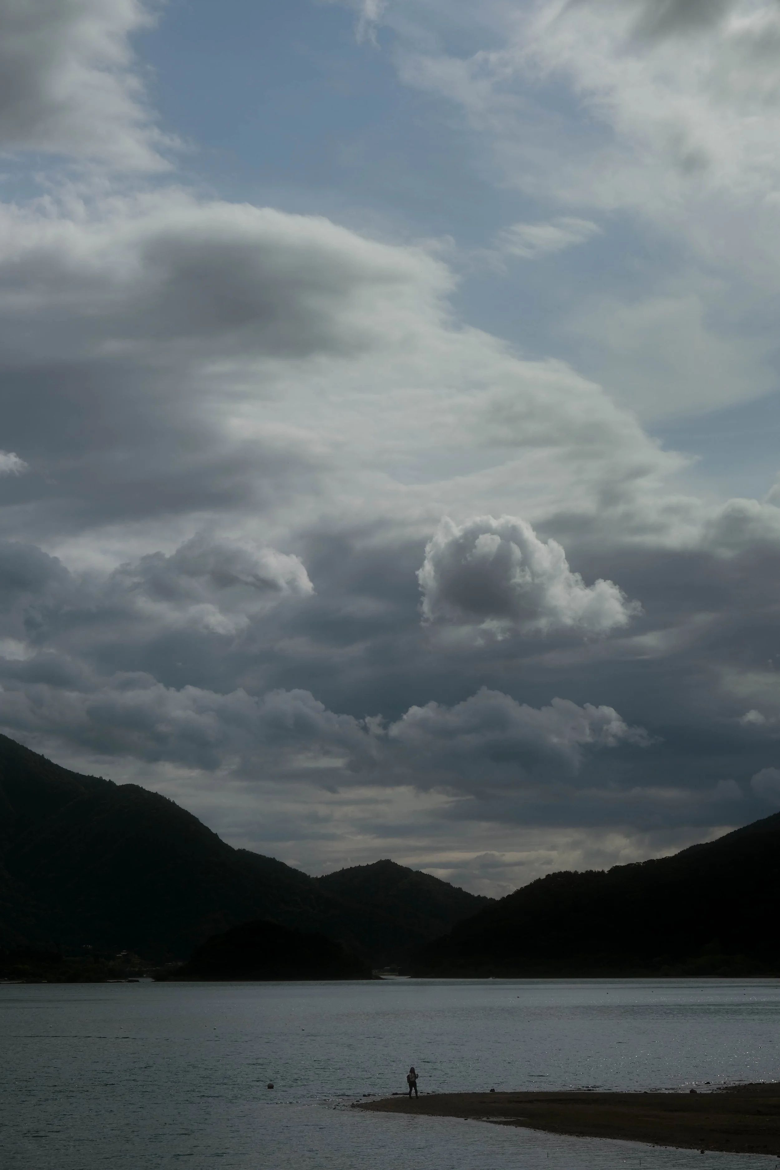 A person standing on a pier in front of a body of water, with mountains and a cloudy sky in the background.