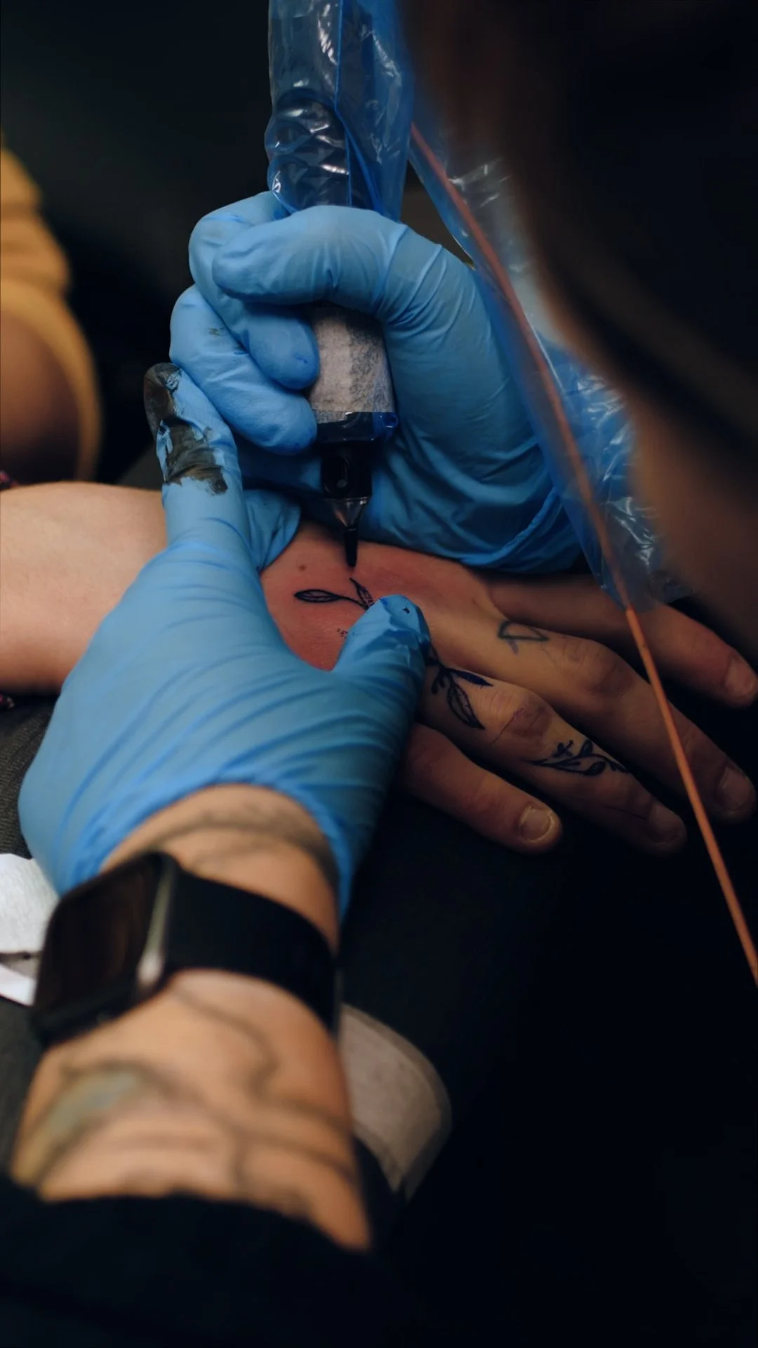 A tattoo artist wearing blue gloves using a tattoo machine to ink a design on a person's hand.