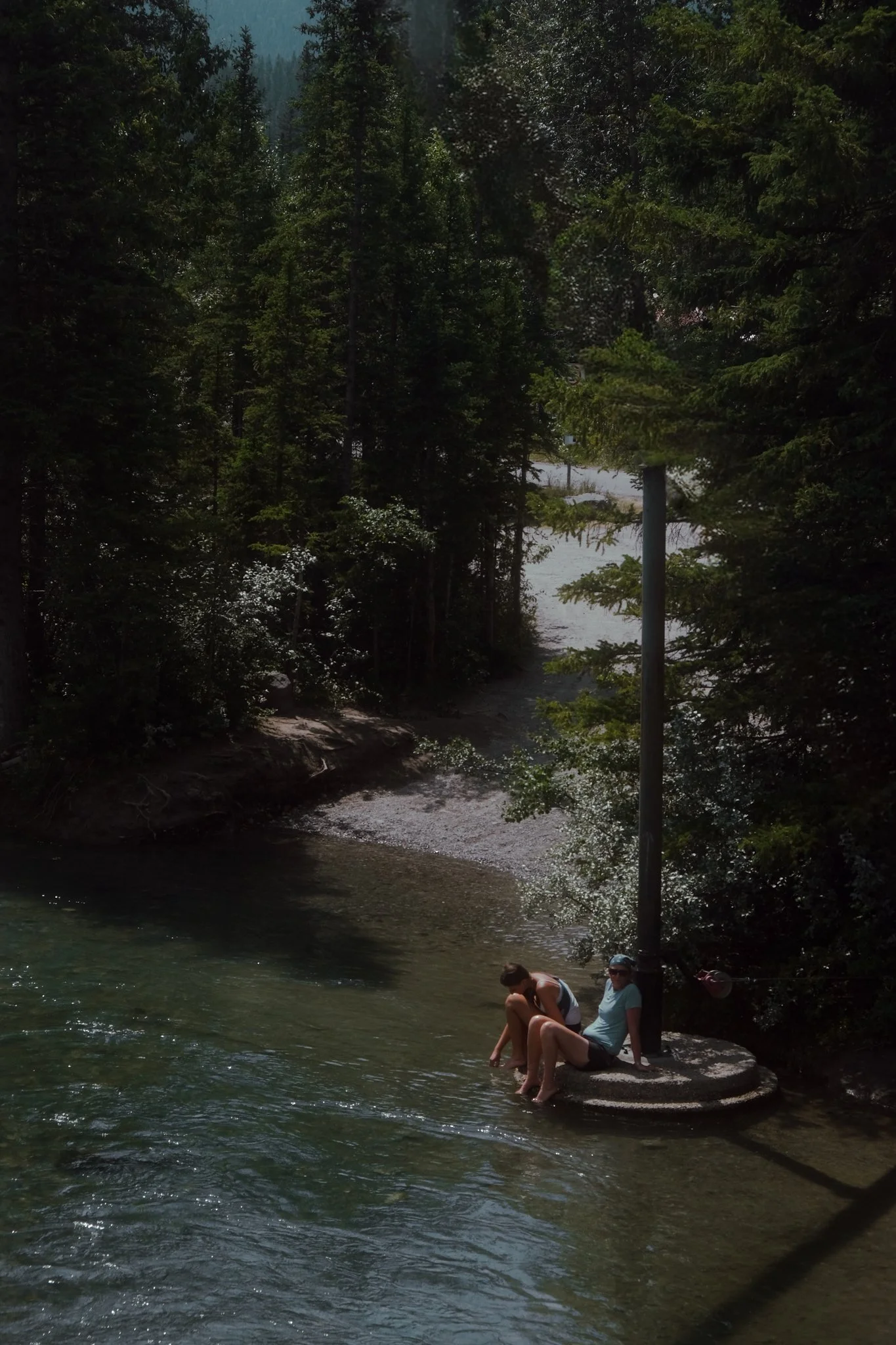People sitting on a rock by a river in a wooded area with tall evergreen trees and mountains in the background.